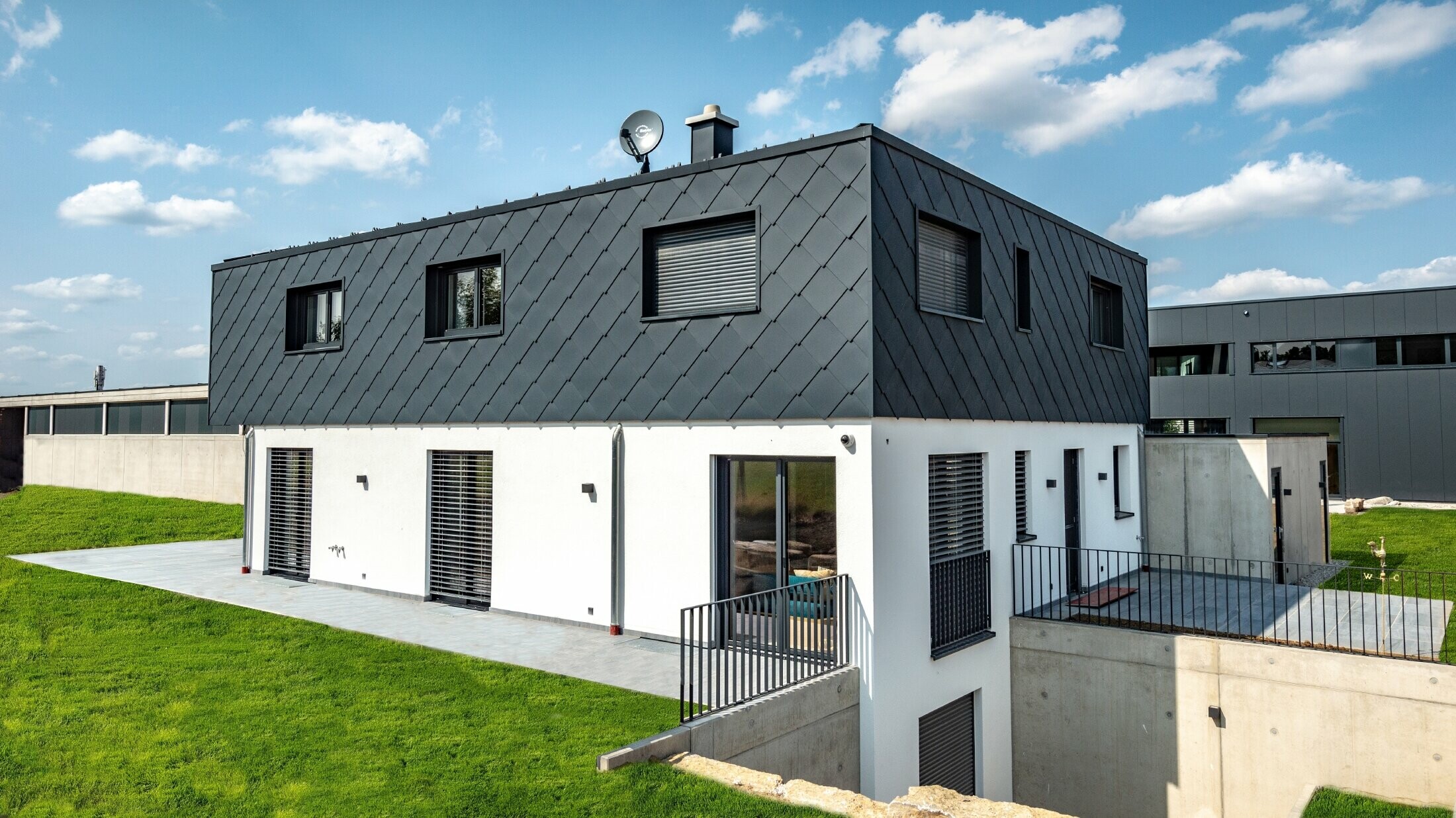 Semplice edificio di nuova costruzione con copertura piatta, il piano terra è intonacato di bianco, il piano superiore è rivestito con la scaglia PREFA in antracite.