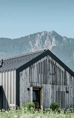 Moderner, funktionaler Wohnbau mit einer Kombination aus Holzfassade und PREFA Band- und Blechmaterial für das Dach. Die klare Architektur wird durch die elegante, metallische Dach- und Fassadenverkleidung unterstrichen, die ideal für anspruchsvolle Projekte ist. Das Bild veranschaulicht die vielseitigen Anwendungsmöglichkeiten der PREFA Produkte im Bereich Bänder und Bleche.