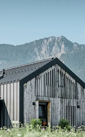 Moderner, funktionaler Wohnbau mit einer Kombination aus Holzfassade und PREFA Band- und Blechmaterial für das Dach. Die klare Architektur wird durch die elegante, metallische Dach- und Fassadenverkleidung unterstrichen, die ideal für anspruchsvolle Projekte ist. Das Bild veranschaulicht die vielseitigen Anwendungsmöglichkeiten der PREFA Produkte im Bereich Bänder und Bleche.