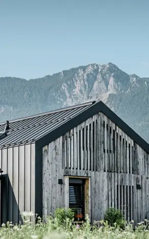 Moderner, funktionaler Wohnbau mit einer Kombination aus Holzfassade und PREFA Band- und Blechmaterial für das Dach. Die klare Architektur wird durch die elegante, metallische Dach- und Fassadenverkleidung unterstrichen, die ideal für anspruchsvolle Projekte ist. Das Bild veranschaulicht die vielseitigen Anwendungsmöglichkeiten der PREFA Produkte im Bereich Bänder und Bleche.