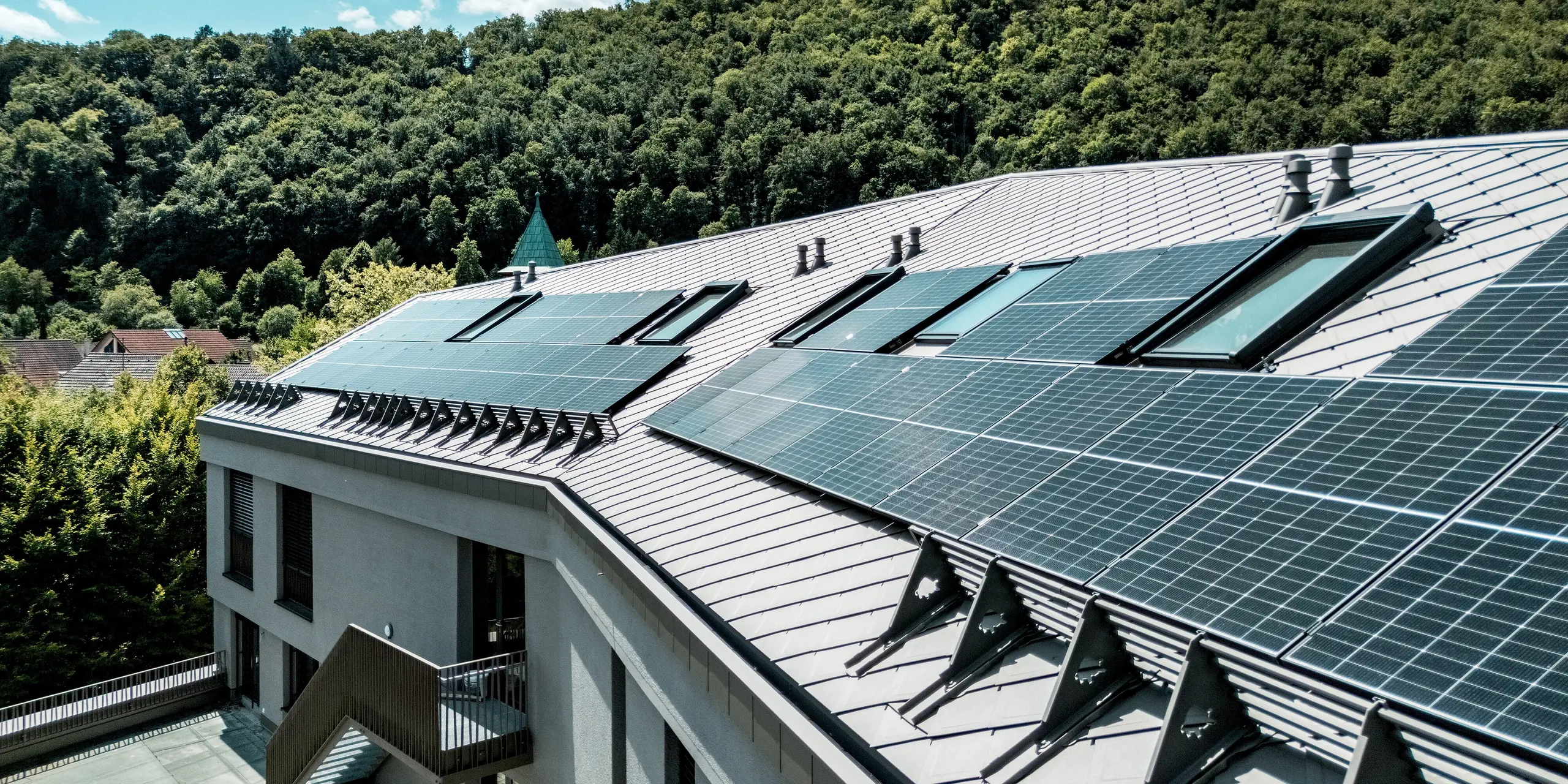 Die Sparkasse Blaubeuren beeindruckt mit ihrer modernen Dachgestaltung aus PREFA Dachrauten 44 × 44 in Bronze. Großflächige Photovoltaikanlagen, integrierte Dachfenster und markante Schneefangsysteme prägen die abwechslungsreiche Dachlandschaft. Die leichten, langlebigen und witterungsbeständigen Aluminiumrauten verbinden elegante Optik mit hoher Funktionalität und fügen sich harmonisch in die grüne Umgebung ein.