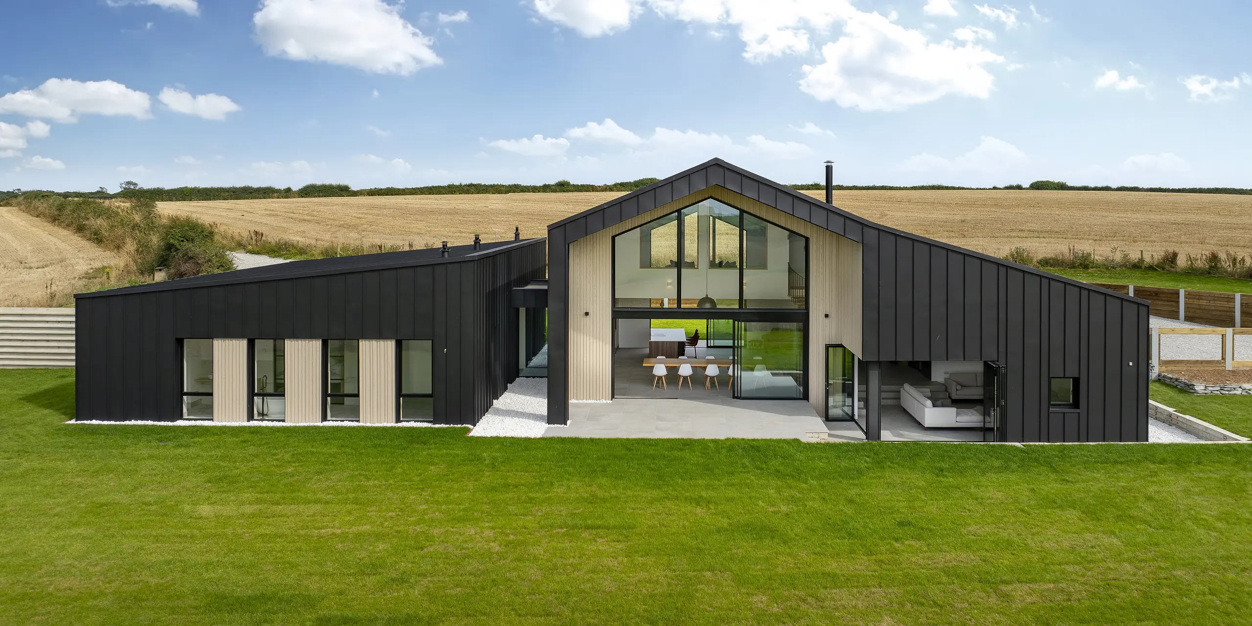 Vue frontale de l'arrière de « The Hide » à Cornwall. La généreuse façade vitrée, encadrée par un revêtement en aluminium PREFALZ en P.10 anthracite, ouvre l'intérieur sur le spacieux jardin. L'aspect clair des joints debout associé à des éléments en bois naturel souligne le design moderne et harmonieux.