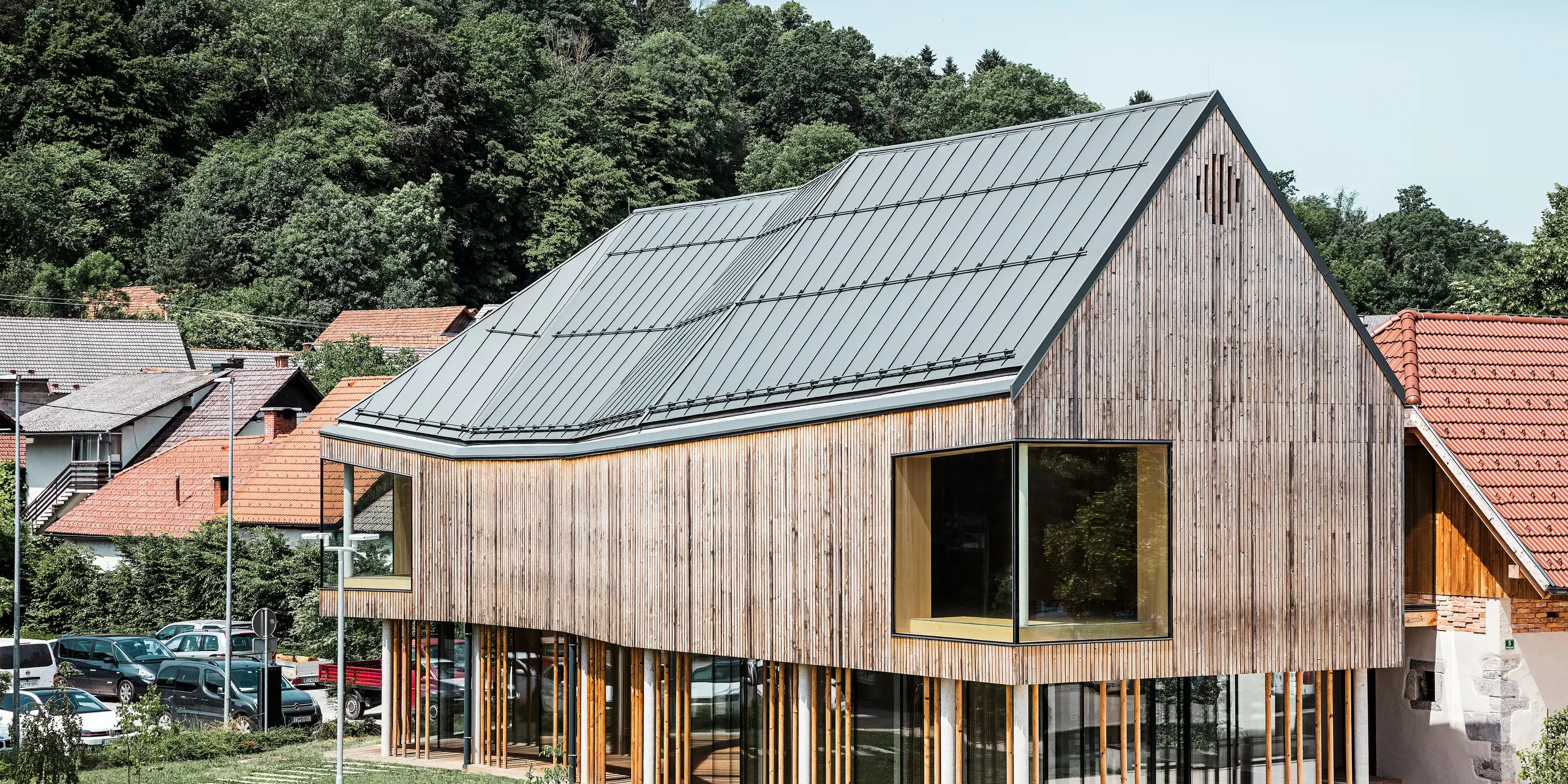 Nahaufnahme des Ig-Besucherzentrums in Slowenien, das sich durch eine moderne Holzfassade auszeichnet und von einer Reihe schlanker Säulen getragen wird. Das charakteristische dunkelgraue Dach von PREFALZ fügt sich nahtlos in die natürliche und ländliche Umgebung ein und bildet einen spannenden Kontrast zu den umliegenden traditionellen roten Ziegeldächern. Dieses Gebäude ist ein Beispiel für das Engagement von PREFA für innovative und nachhaltige Baumaterialien.