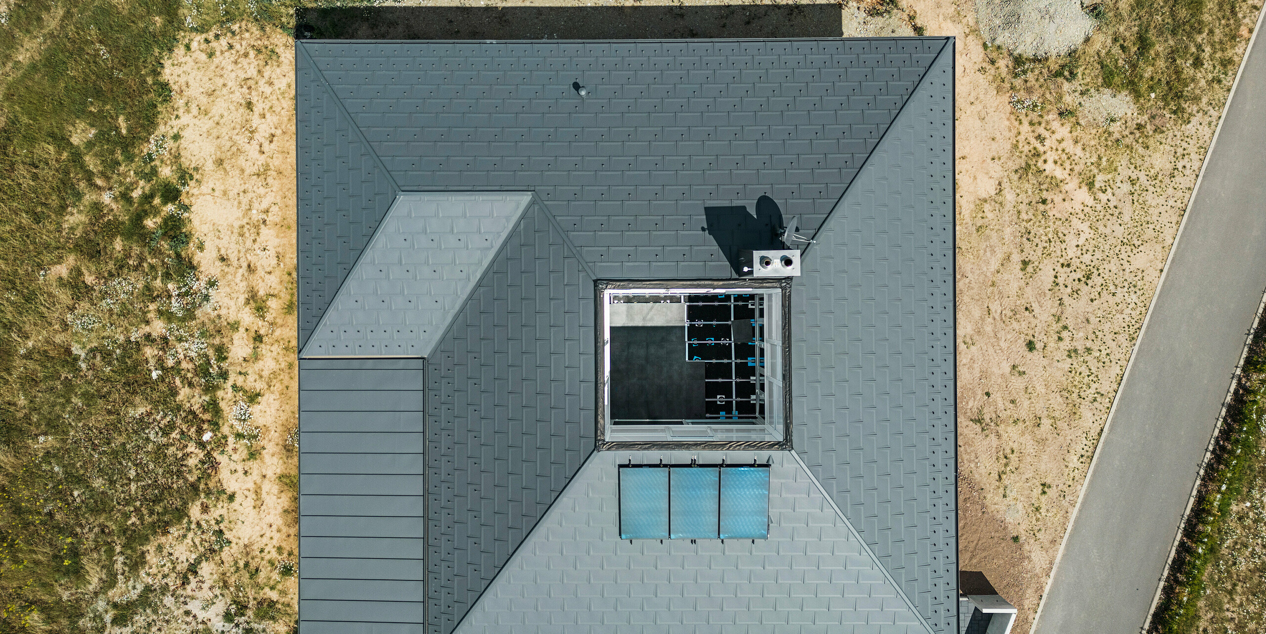 Vogelperspektive eines Einfamilienhauses in Ilmenau, Thüringen, mit einem hochwertigen PREFA Dach aus Dachplatten R.16 und PREFALZ in P.10 Dunkelgrau. Das Bild zeigt die asymmetrische Dachkonstruktion mit integriertem Atrium im Zentrum. Das Atrium setzt nicht nur architektonische Akzente, sondern optimiert auch die Lichtführung im Inneren des Hauses. Die klare Linienführung und die robuste Bauweise des Aluminiumdaches stehen exemplarisch für moderne Dachdesigns und nachhaltige Bauqualität.