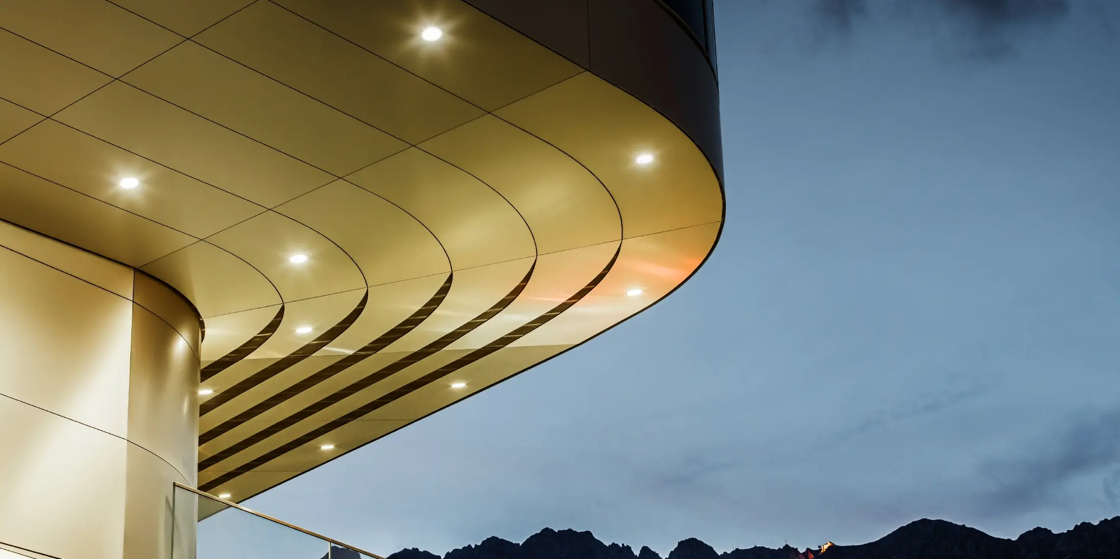Das Bild zeigt eine beeindruckende architektonische Detailsansicht einer PREFA PEMA Verbundplatte aus gebürstetem Aluminium. Die Platte ist sanft gewölbt und verläuft in eleganten, fließenden Linien, die durch gezielte Beleuchtung am Abend besonders zur Geltung kommen. Die klare Struktur und der edle Aluminium-Look der Platte ergänzen das moderne Design der Fassade und bieten eine elegante, zeitgenössische Lösung für hochwertige Architektur.