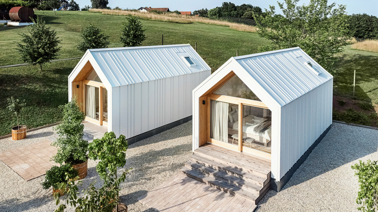 Vue de la façade des deux petites maisons du complexe de vacances « Pri Momi » à Kančevci, en Slovénie. Les micro-maisons ont été construites à ossature bois et dotées d'un revêtement de toit et de façade PREFALZ robuste en blanc pur P.10. Les bâtiments modernes et minimalistes combinent une coque en aluminium à la fois durable et esthétique avec de grandes façades en verre et en bois naturel, intégrés dans un paysage verdoyant. Les spacieuses marches en bois mènent aux entrées, créant une atmosphère chaleureuse et accueillante.