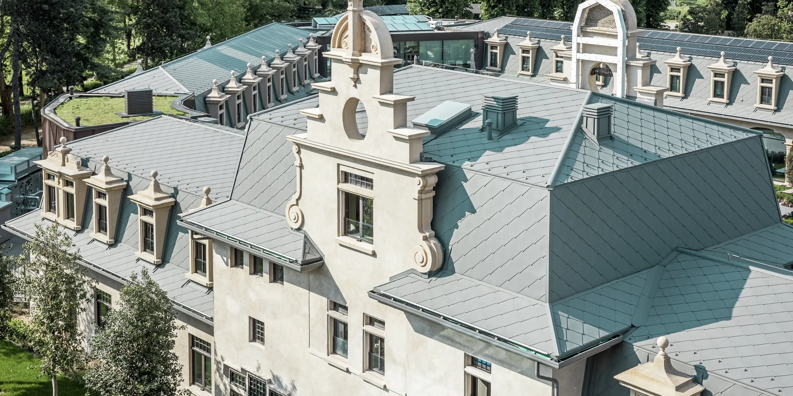 Detaillierte Dachansicht des Thermalparks „De Montel – Terme Milano“ mit präzise ausgeführter Eindeckung aus PREFA Dachrauten 44 × 44 in P.10 Hellgrau. Die komplexe Dachgeometrie mit zahlreichen Gauben, Gesimsen, Giebeln und Lüftungselementen wird handwerklich meisterhaft gelöst. Harmonisch ergänzt wird die historische Architektur durch moderne Elemente wie ein begrüntes Flachdach und die nahtlos integrierte Photovoltaikanlage im Hintergrund.