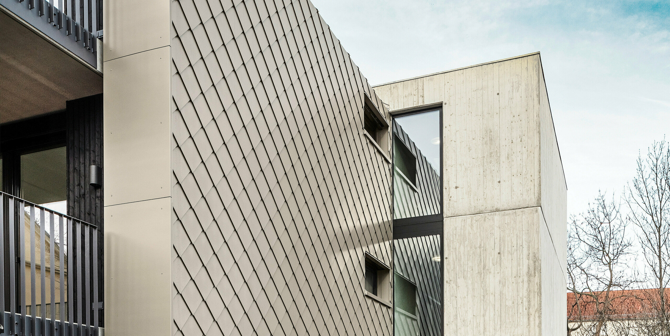 Zone d'accès à un immeuble d'appartements situé dans la Chemnitzer Straße, dans le quartier de Plauen, à Dresde. Le bâtiment est protégé par un système de toiture et de façade robuste PREFA en aluminium. Les losanges de toiture et de façade PREFA de haute qualité 29 × 29 en bronze assurent un aspect élégant et une protection durable. La photo montre la zone d'entrée, flanquée d'une élégante structure en losange et d'un mur en béton. La conception architecturale combine des matériaux robustes avec des lignes épurées et des éléments modernes, donnant au bâtiment un aspect contemporain et élégant.