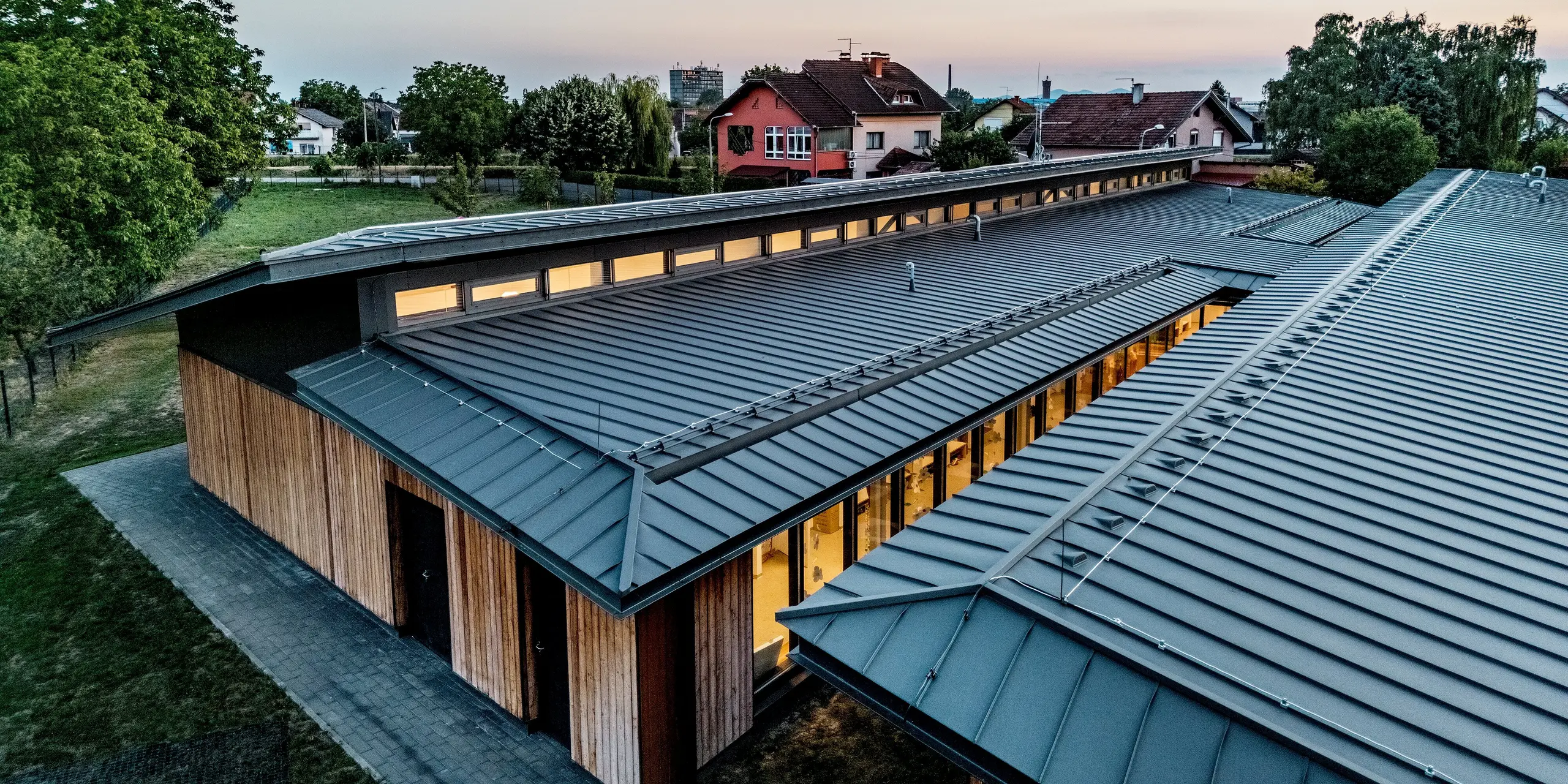 Detailaufnahme des Kindergartens Svarozic mit markanter PREFALZ Dachlandschaft in P.10 Anthrazit. Die präzise gefalzte Aluminiumdeckung betont die klaren Linien des Gebäudes und harmoniert mit der natürlichen Holzfassade. Das moderne Dach mit Oberlichtern und großzügigen Fensterflächen schafft ein helles, einladendes Ambiente und bietet dauerhaften Schutz vor Witterung.