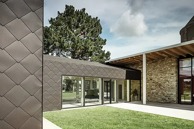 L'immagine mostra una facciata di un edificio moderno rivestita con i scaglie 44 x 44 PREFA per tetto e facciatra nel colore marrone testa di moro. La struttura a diamante del rivestimento in alluminio conferisce alla facciata un aspetto elegante e dinamico. Le ampie vetrate aprono l'interno alla natura circostante, mentre la combinazione di pareti in pietra e rivestimenti moderni crea una transizione armoniosa tra architettura tradizionale e contemporanea. Il prato verde completa l'aspetto naturale e invitante dell'edificio.