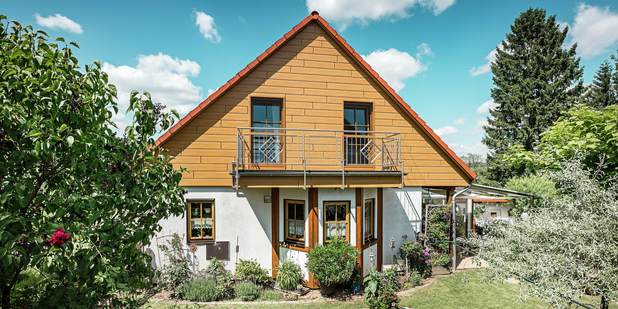 Sur cette image, le pignon rénové d'une maison individuelle à Blieskastel resplendit d'une chaleureuse teinte chêne naturel grâce au Siding PREFA. La façade en aluminium de haute qualité, à l'aspect bois réaliste, crée un contraste saisissant et s'harmonise parfaitement avec le toit de tuiles rouges et le jardin luxuriant. Entourée de fleurs, d'arbustes et de verdure, la maison paraît accueillante et impeccablement entretenue. La surface PREFA allie un aspect naturel à des avantages fonctionnels tels qu'une grande résistance aux intempéries, une stabilité optimale et une durabilité quasi sans entretien – un atout esthétique indéniable et une valeur ajoutée durable.