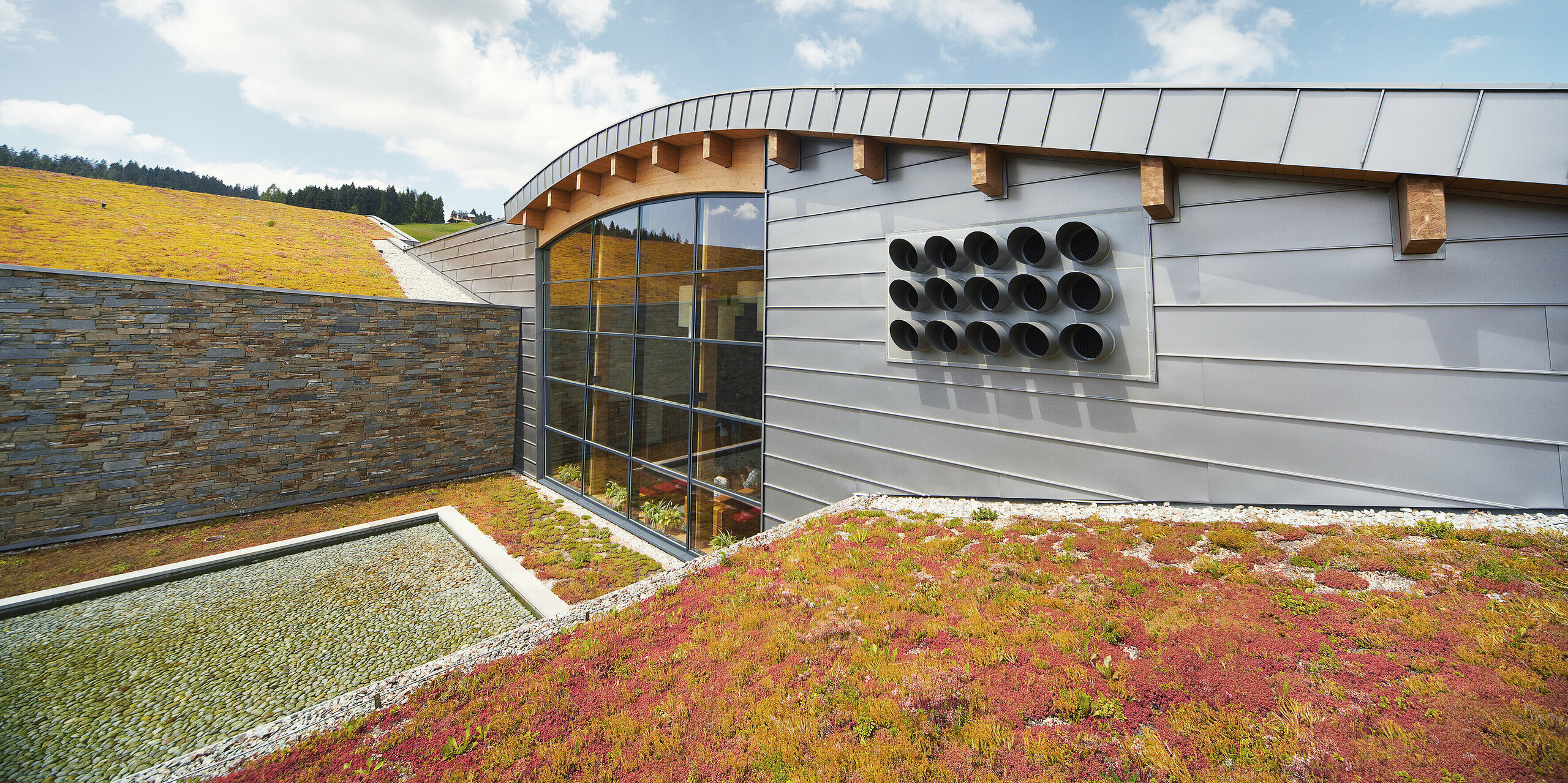 Die Therme Białka im polnischen Tatrzańska besticht durch moderne Architektur und eine faszinierende Fassade aus PREFALZ in der Sonderfarbe Delphin. Der Aluminiumwerkstoff fügt sich perfekt in die Umgebung ein und bietet sowohl ästhetische als auch funktionale Vorteile für das Gebäude. Das harmonische Zusammenspiel von Aluminium, Stein und Grün schafft eine einladende und zeitgemäße Atmosphäre. Die begrünte Dachfläche unterstreicht den ökologischen Ansatz der Architektur.
