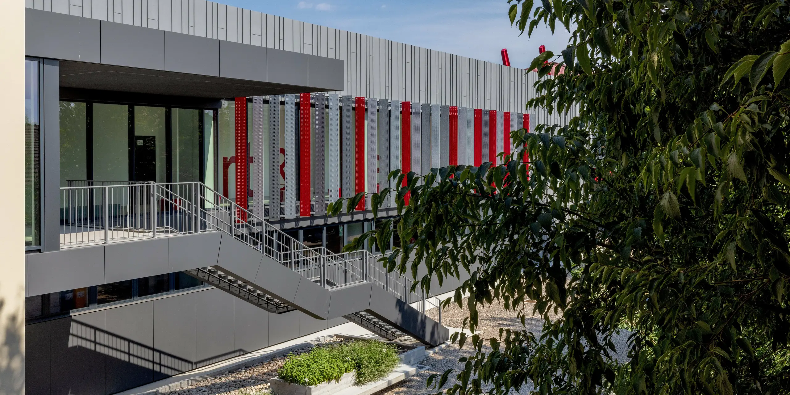 L'espace extérieur animé de la Scuola Arte e Mestieri F. Bertazzoni à Suzzara est agrémenté d'un escalier extérieur longeant la façade en aluminium PREFA, de teinte gris sombre et gris quartz, et menant au niveau d'entrée. Des bandes rouges disposées de façon rythmée et des éléments perforés confèrent au bâtiment une énergie dynamique. À l'intérieur, une grande inscription rouge souligne le caractère distinctif de l'établissement. Les produits PREFALZ et PREFABOND de PREFA impressionnent par leurs finitions impeccables, leur stabilité dimensionnelle exceptionnelle et leur résistance durable aux intempéries. Ils s'intègrent harmonieusement dans un espace vert accueillant au charme méditerranéen.