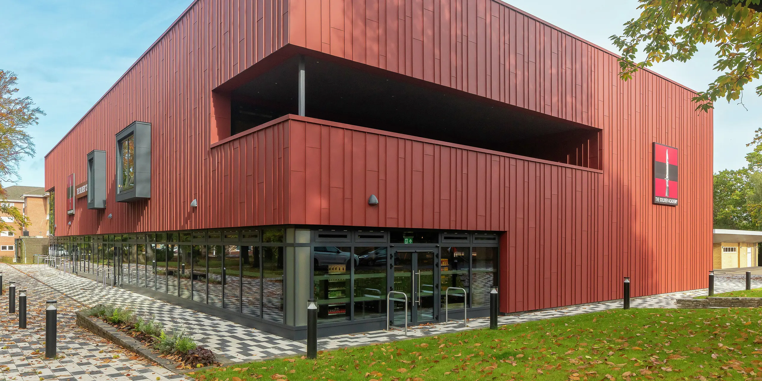 Architecture contemporaine avec PREFALZ : Le Soldiers' Centre de Pirbright impressionne par sa façade moderne à joint debout en PREFALZ en P.10 rouge oxyde. Les vastes baies vitrées du rez-de-chaussée créent transparence et ouverture, tandis que les cadres de fenêtres en P.10 anthracite ajoutent des accents élégants. Un concept de construction durable avec des produits en aluminium PREFA durables pour les normes de conception et de qualité les plus élevées.