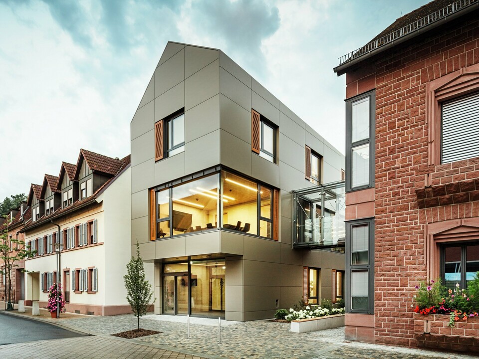 Das Bild zeigt das moderne Rathaus in Alzenau, dessen Fassade mit PREFA-Aluminiumverbundplatten in einem warmen Bronzeton verkleidet ist. Große Glasflächen unterbrechen die Fassade und geben den Blick auf ein lichtdurchflutetes Interieur frei. Ein gläserner Verbindungsgang auf der rechten Bildseite verbindet das Gebäude mit einem nahegelegenen traditionellen Backsteingebäude und schafft so einen interessanten Kontrast zwischen moderner und historischer Architektur.