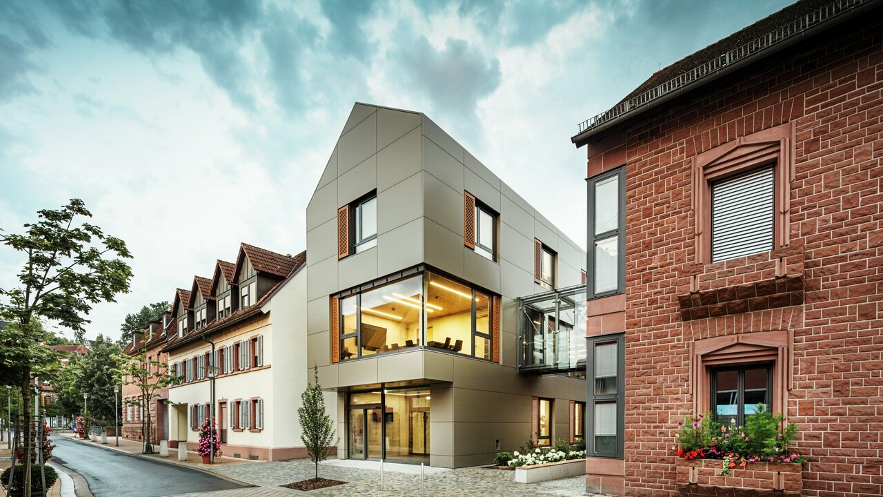 La photo montre la mairie moderne d'Alzenau, revêtue de panneaux composites en aluminium PREFA dans un ton bronze chaleureux. La façade est interrompue par de grandes surfaces vitrées qui offrent une vue sur l'intérieur, éclairé par une lumière chaude. La passerelle vitrée à droite de l'image relie le bâtiment à un bâtiment traditionnel en briques situé à proximité, créant ainsi un contraste intéressant entre l'architecture moderne et historique.