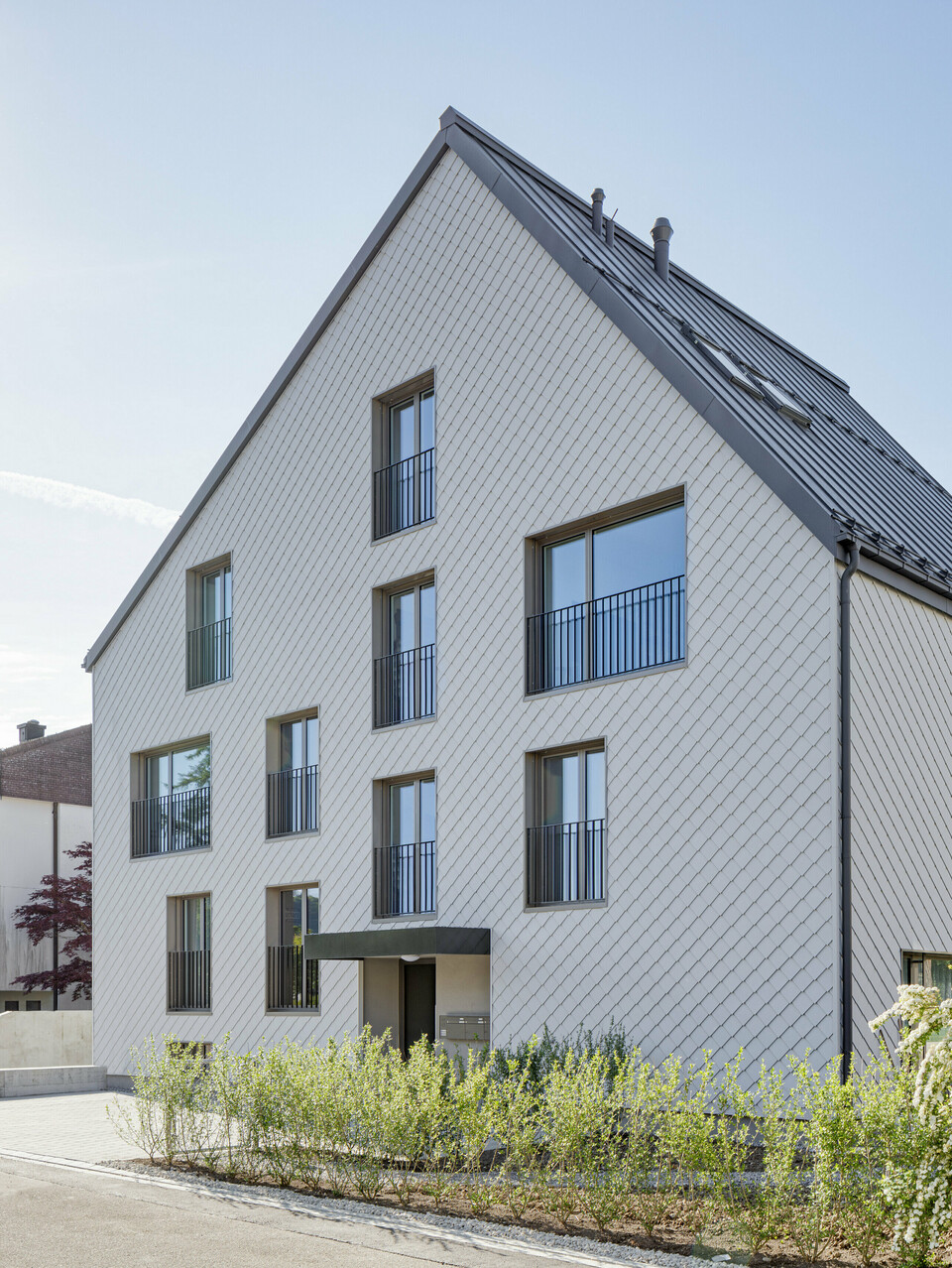 Modernes Mehrfamilienhaus in der Schweiz mit einem langlebigen PREFA Dach aus PREFALZ in P.10 Dunkelgrau und einer Fassade, verkleidet mit PREFA Wandrauten 20x20 in P.10 Prefaweiß. Das Objekt bietet eine zeitgemäße Architektur, die durch die Kombination von traditionellem Satteldach und minimalistischen Fassadenelementen besticht. Die klare Linienführung und das strukturierte Erscheinungsbild der Wandrauten sind harmonisch in eine gepflegte Umgebung mit üppigem Blütenstrauch integriert und zeigen die Vielseitigkeit von Aluminium als Baumaterial.