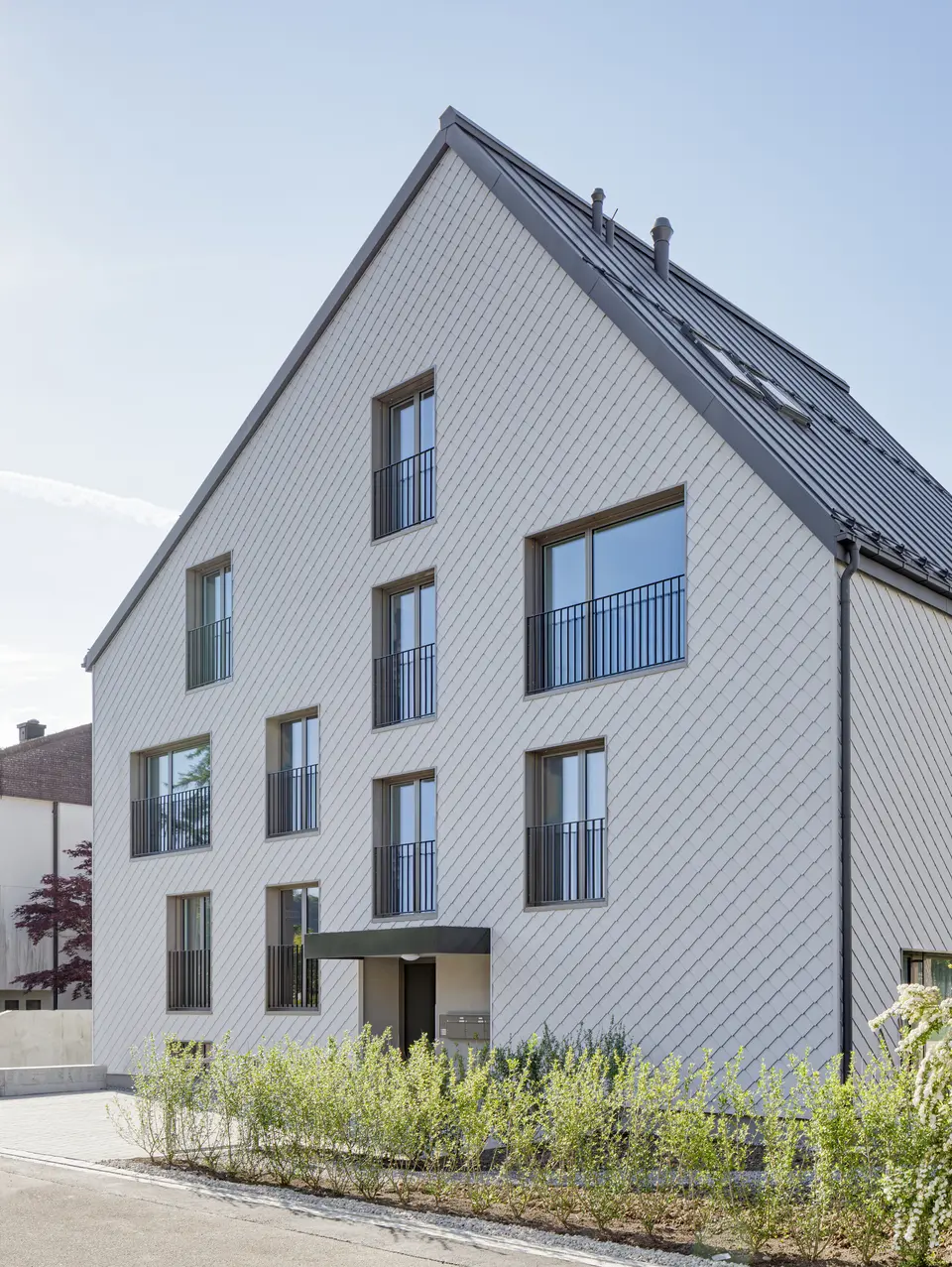 Modernes Mehrfamilienhaus in der Schweiz mit einem langlebigen PREFA Dach aus PREFALZ in P.10 Dunkelgrau und einer Fassade, verkleidet mit PREFA Wandrauten 20x20 in P.10 Prefaweiß. Das Objekt bietet eine zeitgemäße Architektur, die durch die Kombination von traditionellem Satteldach und minimalistischen Fassadenelementen besticht. Die klare Linienführung und das strukturierte Erscheinungsbild der Wandrauten sind harmonisch in eine gepflegte Umgebung mit üppigem Blütenstrauch integriert und zeigen die Vielseitigkeit von Aluminium als Baumaterial.