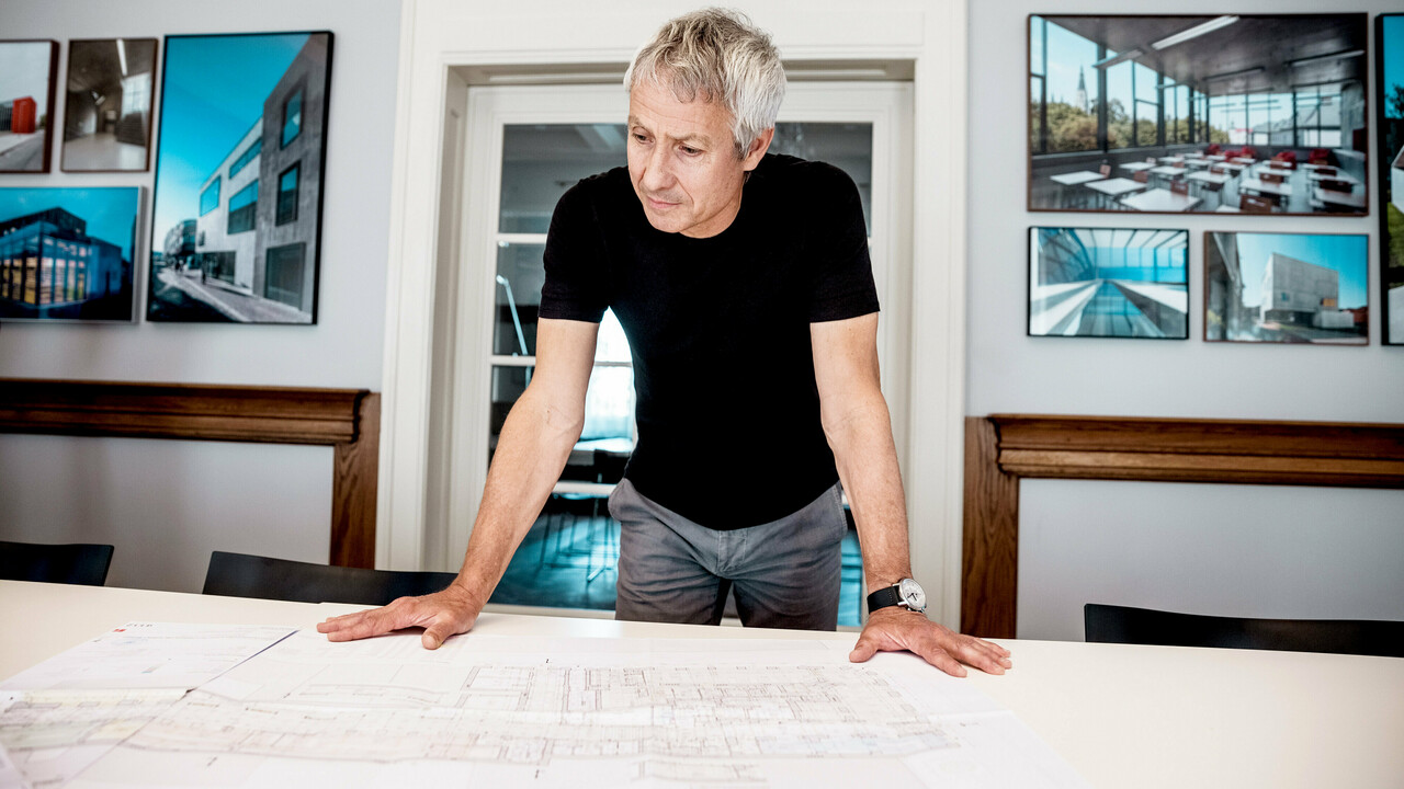 Architekt arbeitet an detaillierten Bauplänen in einem modernen Büro, umgeben von beeindruckenden Fotos von PREFA-Architekturprojekten.