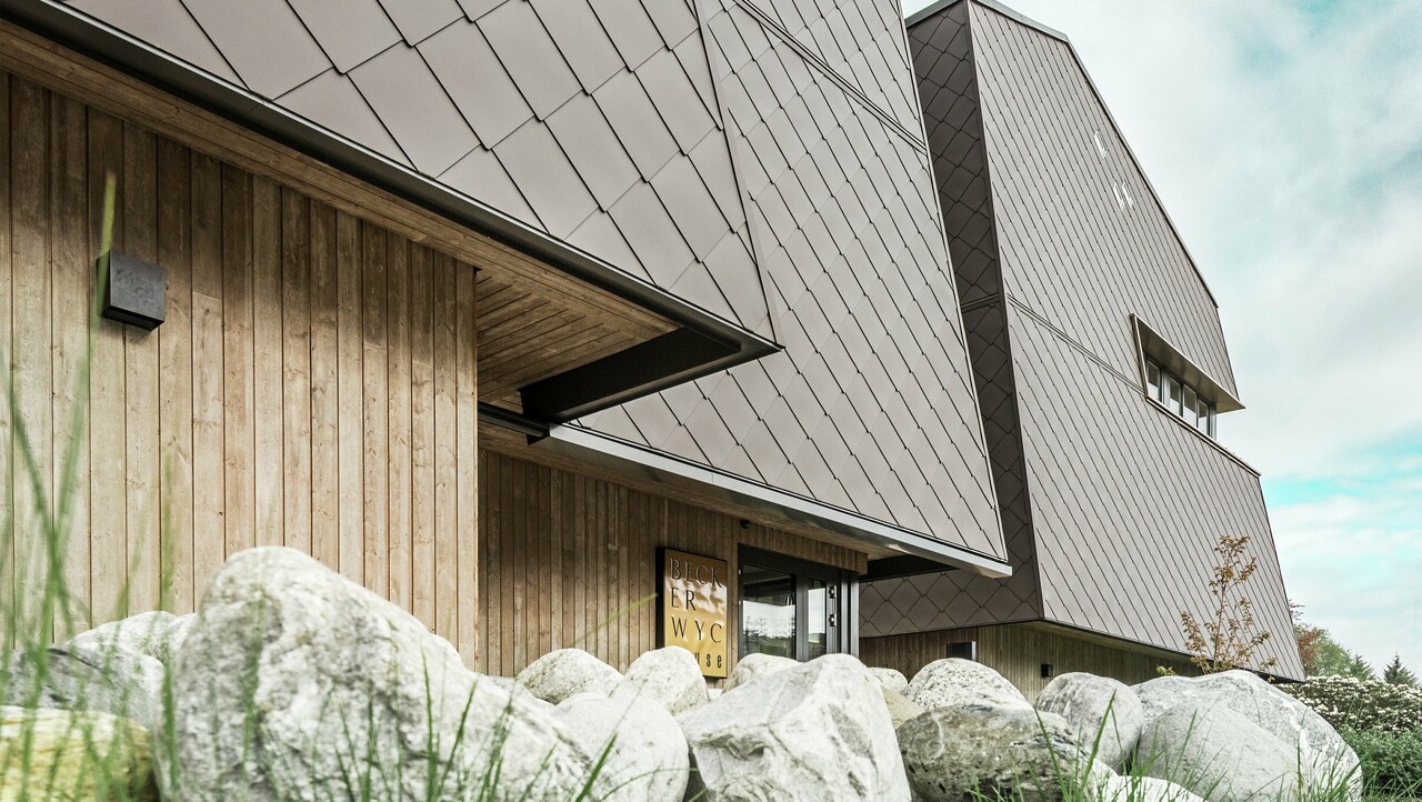 Vista laterale di un hotel, la facciata del piano terra è rivestita verticalmente con legno piuttosto scuro. Sopra c'è una sporgenza della facciata, da qui in poi la facciata è rivestita con pannelli di alluminio color noce.