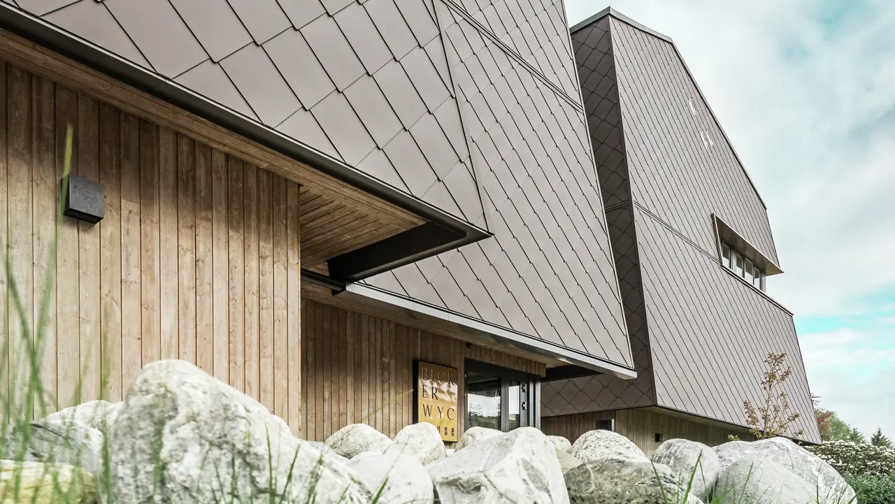 Vue latérale d'un hôtel, la façade du rez-de-chaussée est revêtue verticalement de bois plutôt sombre. Au-dessus se trouve un avant-toit, à partir duquel la façade est revêtue de plaques d'aluminium brun noisette.