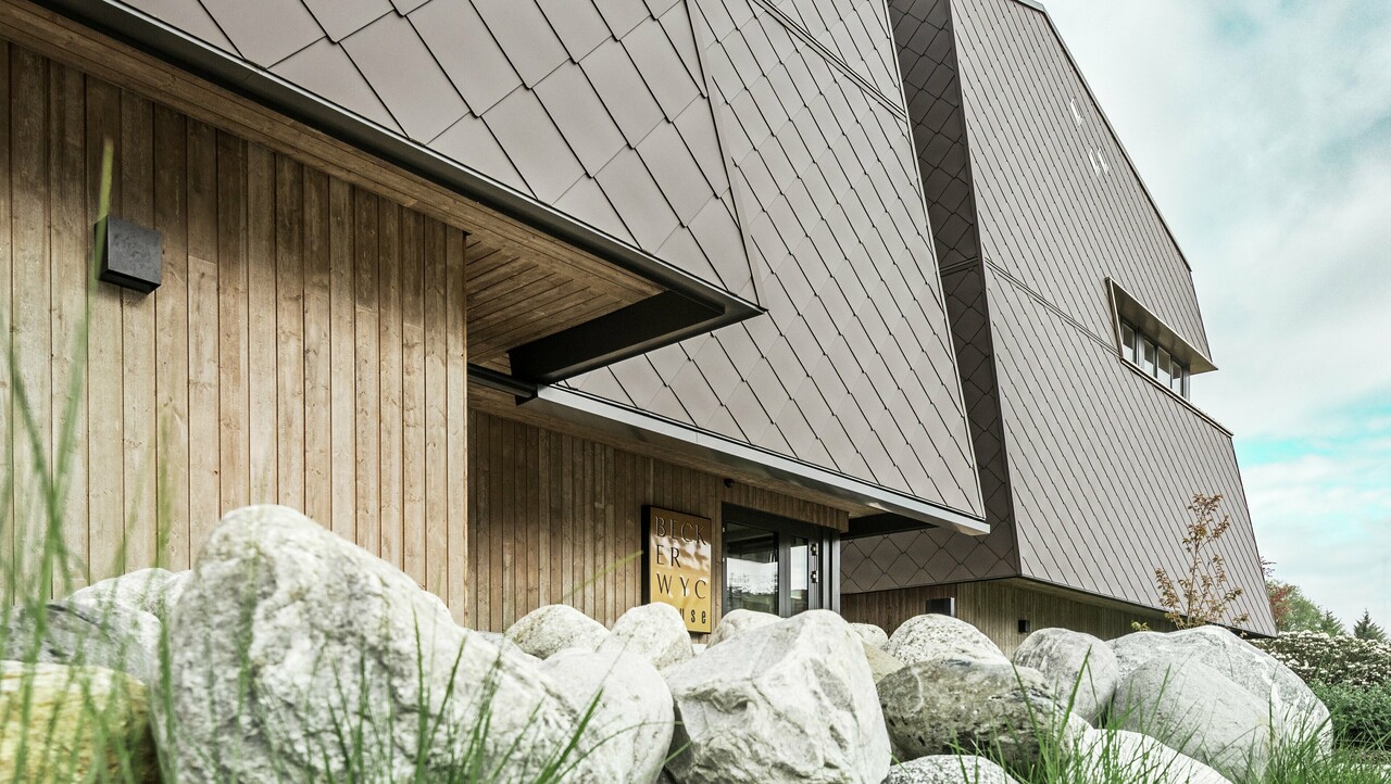 Seitliche Ansicht eines Hotels, die Fassade des Erdgeschosses ist mit eher dunklem Holz vertikal verkleidet. Darüber ist ein Fassadenvorsprung, ab hier ist die Fassade mit Aluminiumplatten in Nussbraun verkleidet.