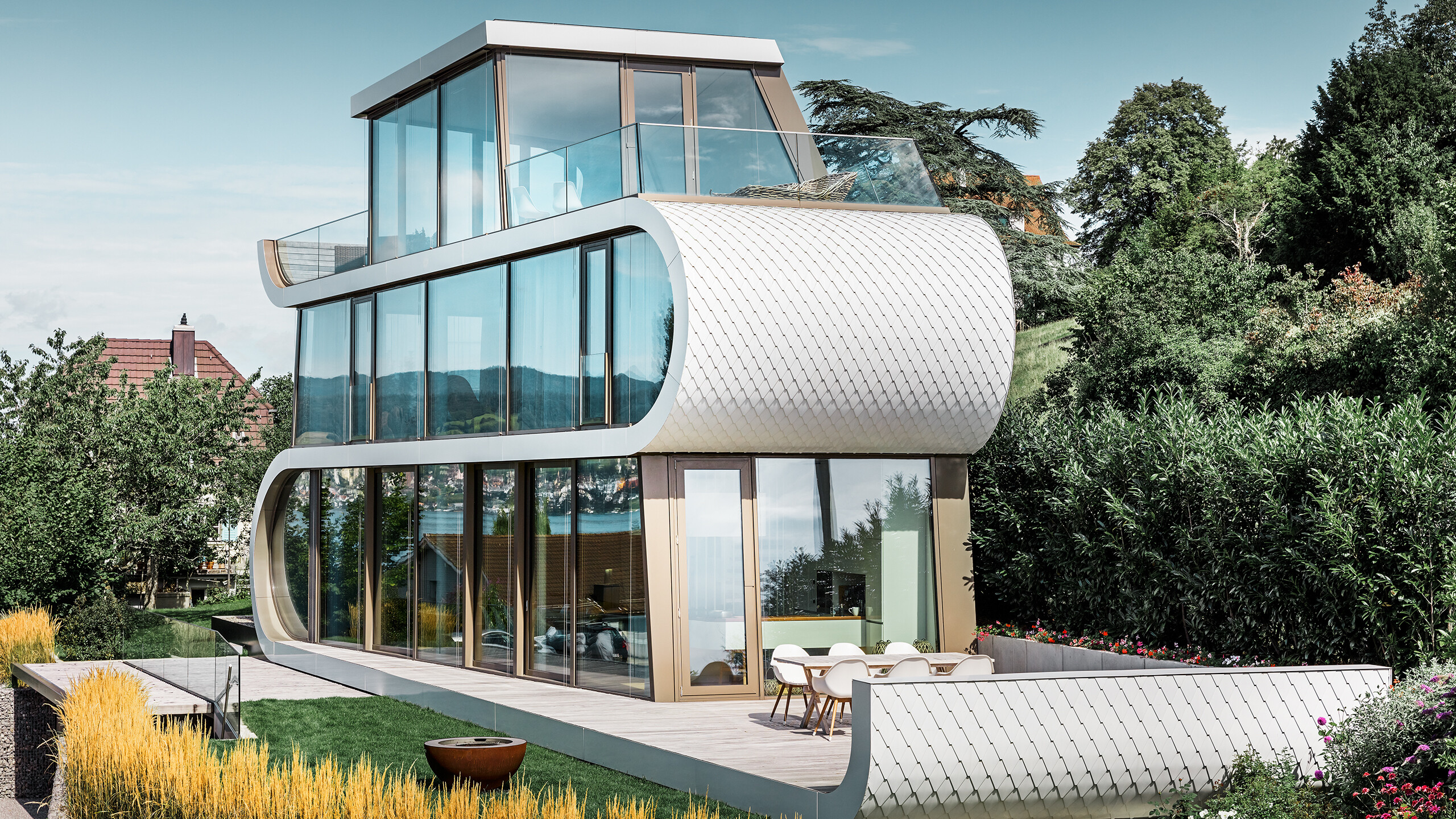 Photo de la Flexhouse moderne au bord du lac de Zurich. Avec sa forme hors du commun, la Flexhouse attire toujours le regard. Les séparations entre étages s’étirent comme une bande blanche ondulée à travers le bâtiment. Les éléments de façade arrondis ont été recouverts de bardeaux PREFA en aluminium blanc pur.