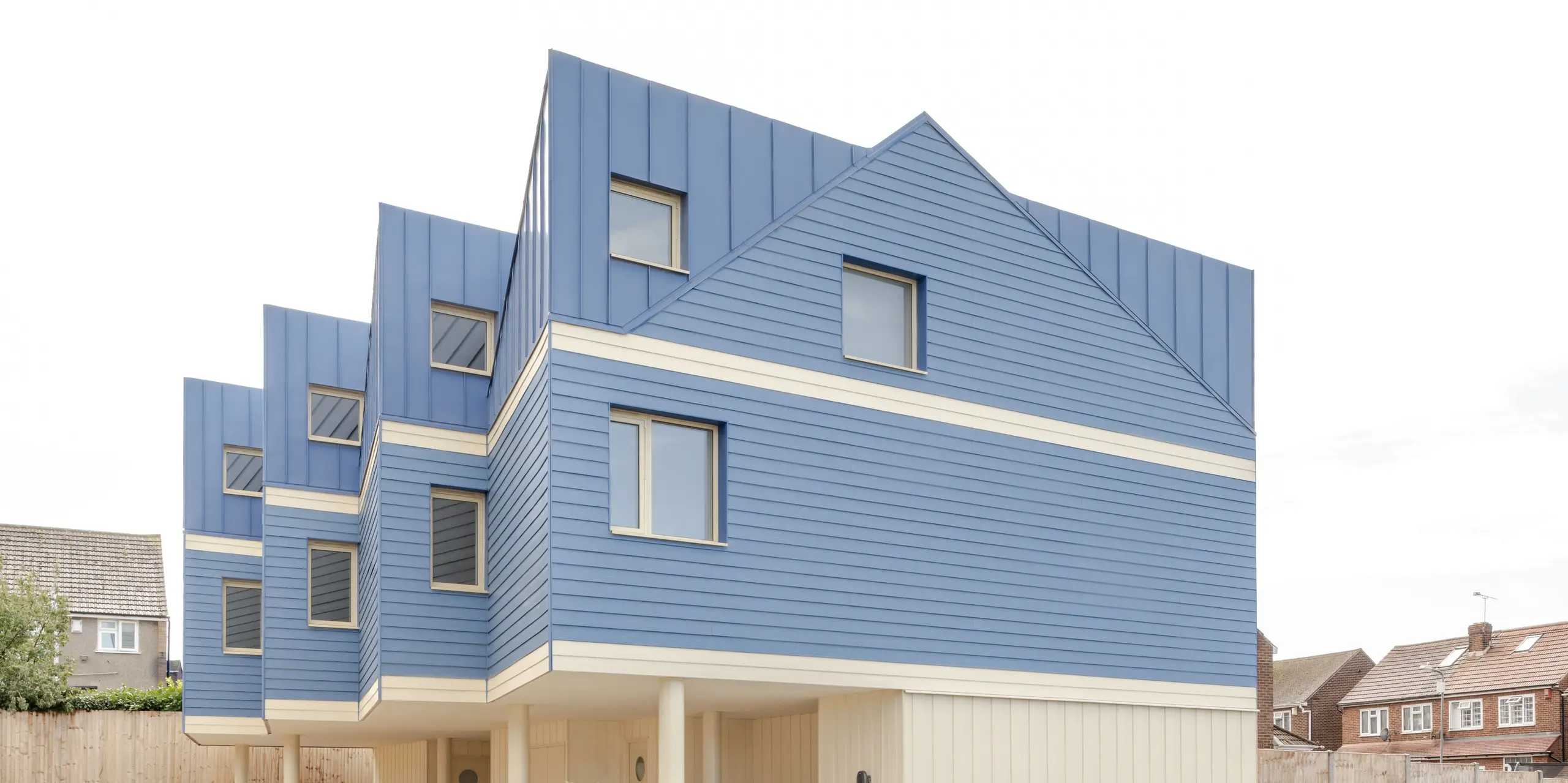 L'image révèle la façade remarquable du complexe résidentiel Goldsmith-Mews, avec son bardage FALZONAL bleu pigeon du plus bel effet. Les panneaux à joints debout verticaux et précis de la toiture et des étages supérieurs s'intègrent harmonieusement aux profilés horizontaux en aluminium, créant une façade dynamique et rythmée. Les parties inférieures claires et la construction surélevée confèrent au bâtiment une impression de légèreté. L'aluminium PREFA, durable et inaltérable, offre une protection fiable contre les intempéries, une grande stabilité dimensionnelle et permet un style architectural moderne et épuré, gage d'une forte identité visuelle.