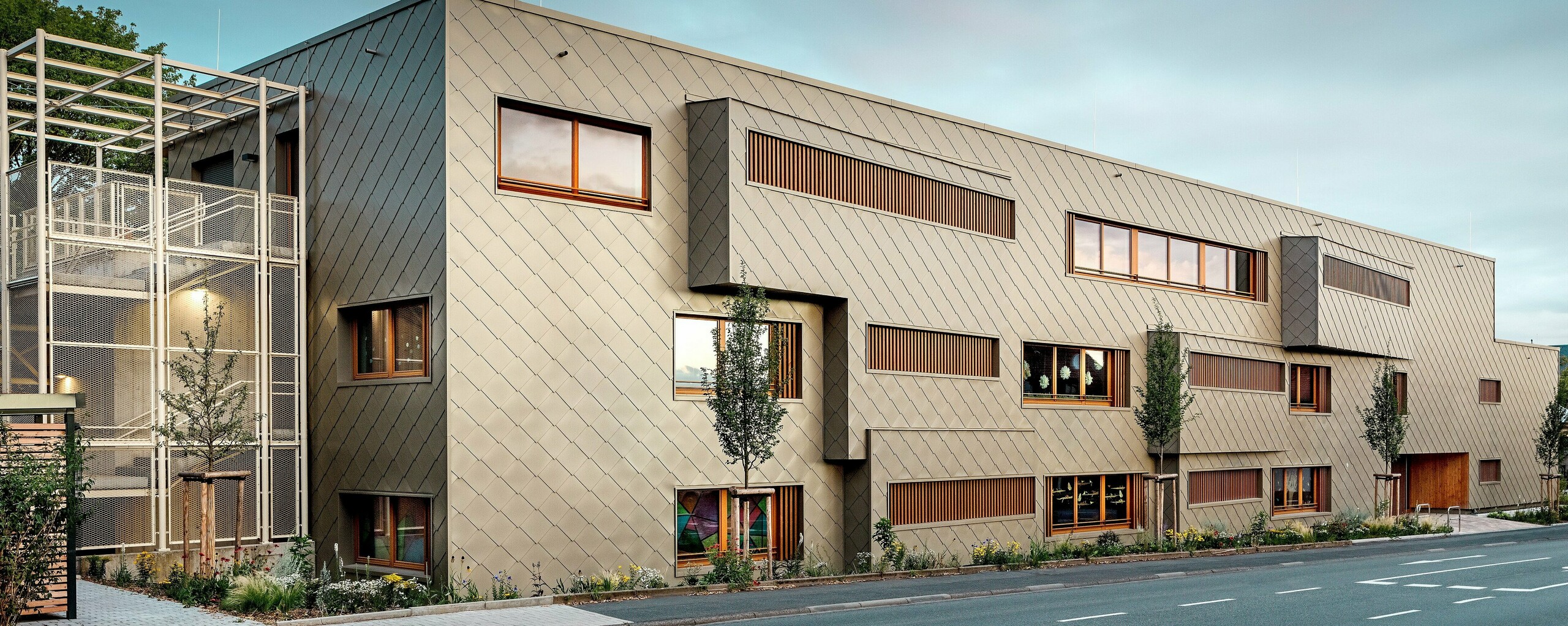 Die Aufnahme aus Straßenniveau zeigt die neue Kindertagesstätte in Alzenau im spannungsvollen Dialog mit der historischen Stadtkulisse, darunter die markante Kirche im Hintergrund. Die moderne Fassade aus bronzefarbenen PREFA Wandrauten 44 × 44 verleiht dem Gebäude eine elegante, fast skulpturale Wirkung. Präzise gesetzte Fenster, vertikale Holzlamellen und versetzte Fassadenflächen schaffen Tiefe und Rhythmus. Links ist das filigrane Treppenhaus mit weißem Metallgerüst sichtbar, das moderne Leichtigkeit vermittelt. Die klare Formsprache, nachhaltige Materialwahl und die hochwertige Aluminiumfassade machen das Gebäude zu einem Vorzeigeprojekt für städtebauliche Integration und zukunftsorientiertes Bauen mit PREFA Systemen.
