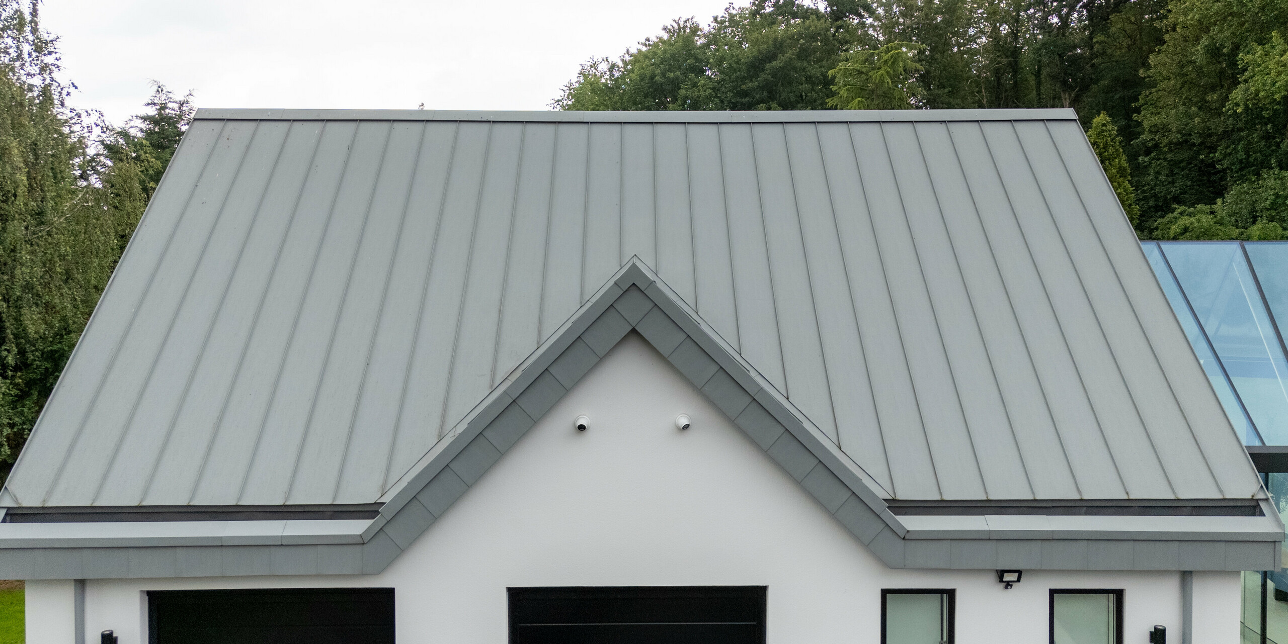 Cette image présente une vue détaillée de la toiture pointue en aluminium PREFALZ, gris quartz, aux joints debout apparents qui soulignent le design moderne de la villa. Sa forme symétrique et sa surface métallique sans joint apparent lui confèrent non seulement une valeur esthétique, mais aussi une grande durabilité et une excellente résistance aux intempéries. L'intégration harmonieuse de la toiture à l'architecture générale du bâtiment, qui se fond parfaitement dans le paysage verdoyant de Draycott In The Clay, renforce l'alliance parfaite de fonctionnalité et de style.