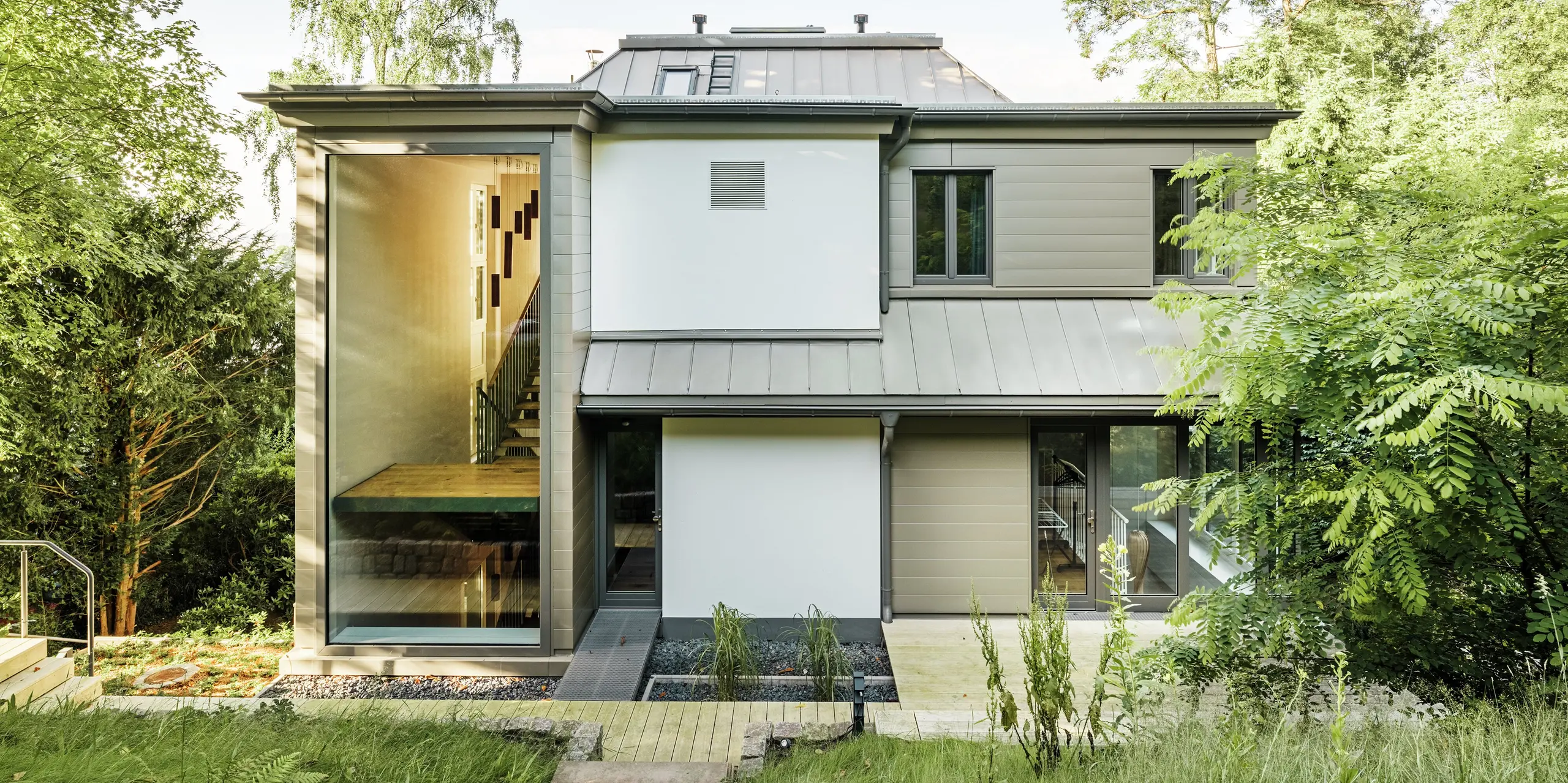 Das Bild zeigt die Rückseite der Villa Blankenese in Hamburg mit einer modernen, klar strukturierten Architektur. Besonders auffällig sind die bronzefarbenen PREFA Sidings, die an der Fassade für eine elegante und zeitlose Optik sorgen. Das Dach ist mit PREFALZ in Bronze ausgeführt und fügt sich harmonisch in das Gesamtbild ein. Große Glasflächen und die Kombination aus Aluminium und Putz unterstreichen den hochwertigen Charakter des Gebäudes. Die langlebigen PREFA Produkte bieten nicht nur ein edles Design, sondern auch maximale Witterungsbeständigkeit und Wartungsfreiheit.