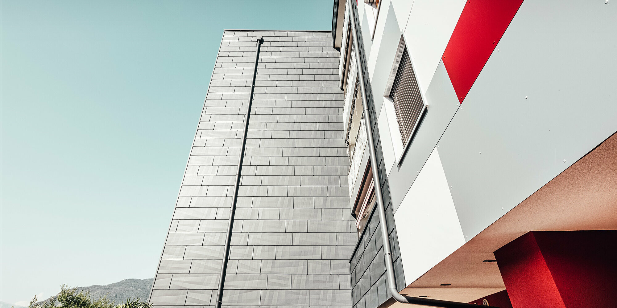 Prise de vue rapprochée et du dessous. En premier plan : la façade PREFA anthracite. A droite : le front de façade dans les teintes blanc, gris et rouge.