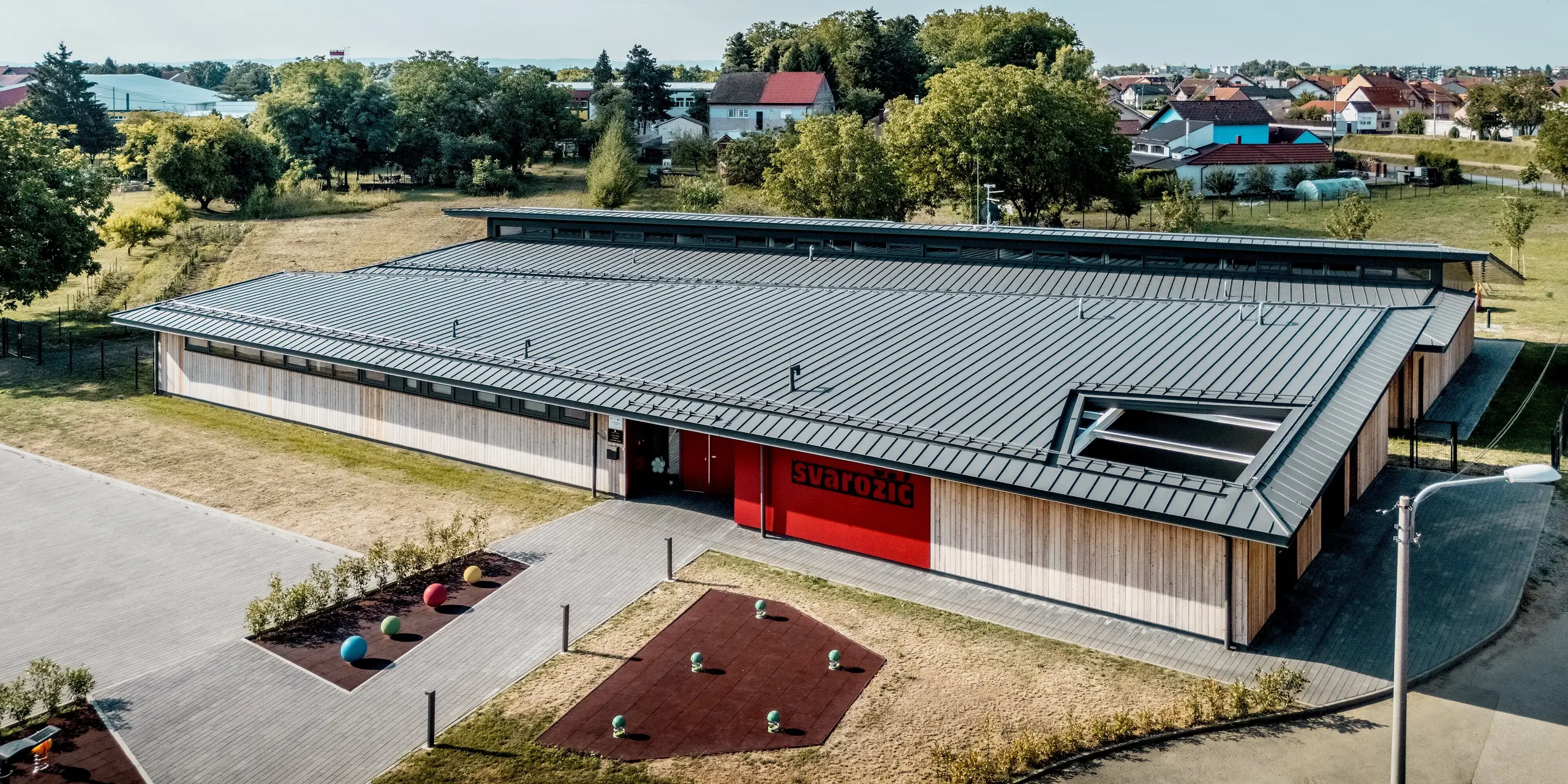Weitläufige Außenansicht des Kindergartens Svarozic in Kroatien mit prägendem PREFALZ Aluminiumdach in P.10 Anthrazit. Die klare Dachsilhouette, natürliche Holzfassade und großzügigen Fensterbänder fügen sich harmonisch in die grüne Umgebung ein. Das PREFA Dach überzeugt durch langlebigen Schutz, hohe Widerstandsfähigkeit und eine elegante, zeitlose Gestaltung.