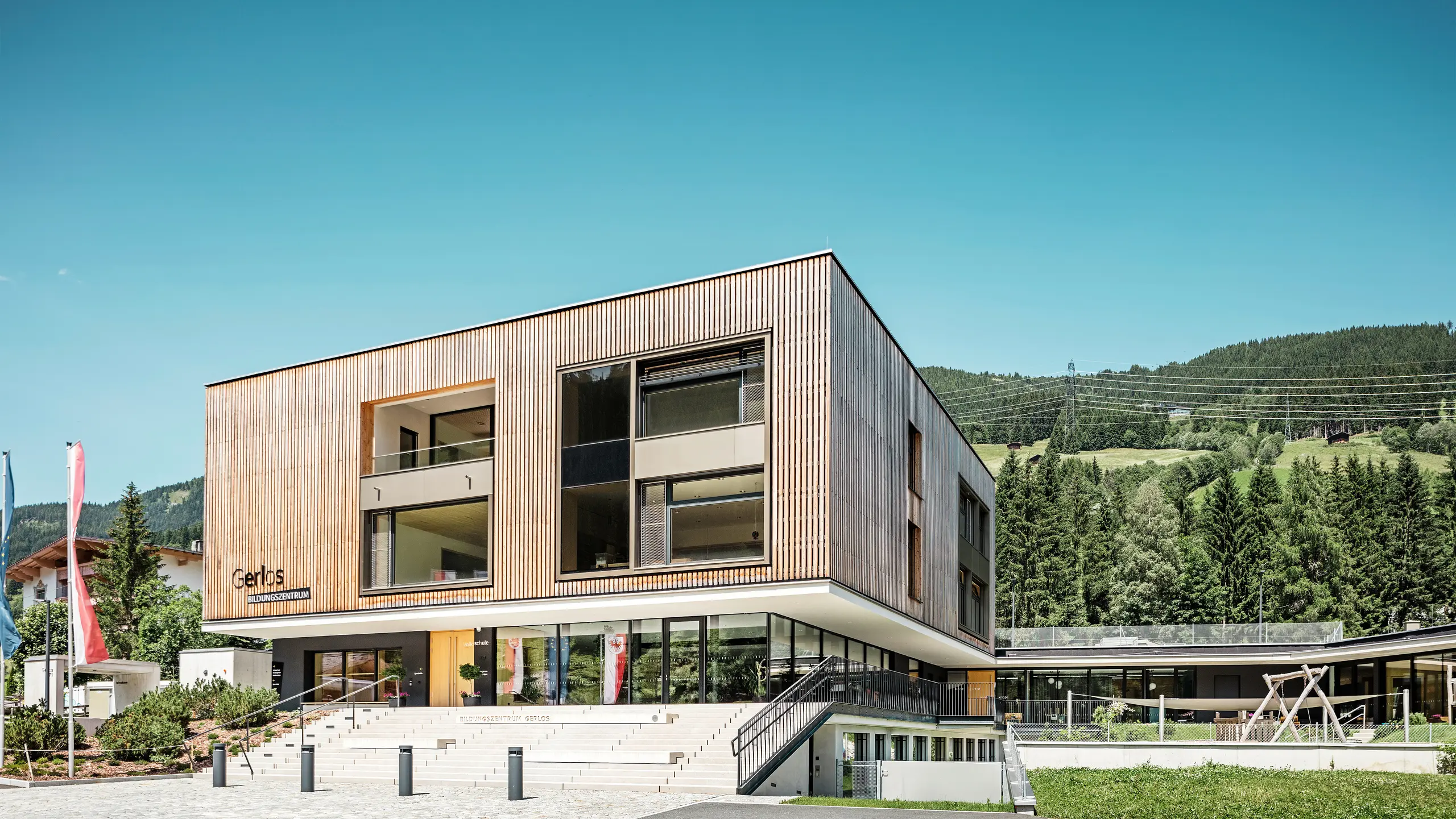 Vista frontale del moderno centro educativo con facciata in legno.  L'edificio è a due piani, con ampie finestre frontali e un tetto piatto che si inserisce perfettamente nel paesaggio montano circostante, caratterizzato da colline densamente boscose. In primo piano ci sono spazi verdi curati, un vialetto a gradini e bandiere pubblicitarie. Il progetto trasmette una combinazione di funzionalità ed estetica architettonica in armonia con la natura. 