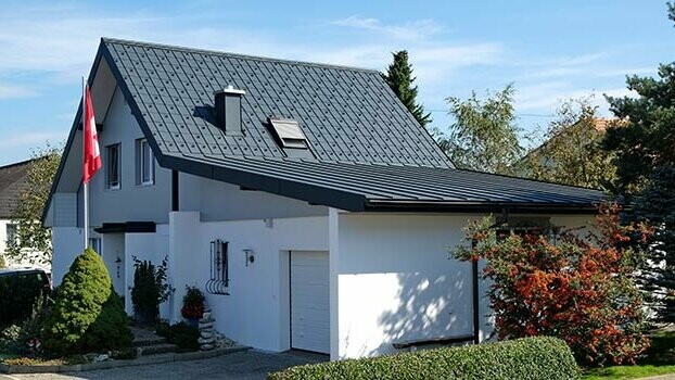 Saniertes Haus mit Satteldach und anschließender Garage. Das Dach wurde mit der PREFA Dachplatte und die Garage mit Prefalz in Anthrazit eingedeckt. Vor dem Haus ist ein Fahnenmast mit Schweiz-Fahne.
