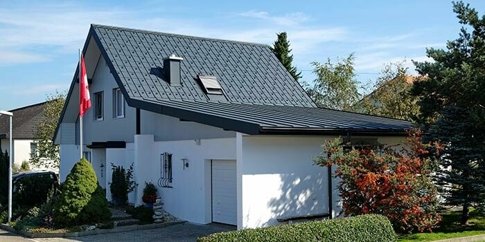 Maison rénovée avec toit à deux pans et garage accolé. Le toit est recouvert de tuiles PREFA et le garage à l’aide de Prefalz couleur anthracite. Un mât avec le drapeau suisse se trouve devant la maison.