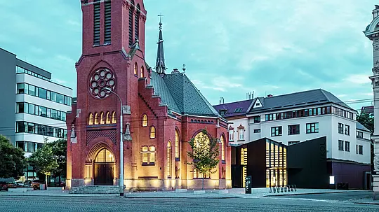 L'immagine mostra l'armoniosa fusione tra architettura storica e moderna a Olomouc. L'imponente Chiesa Rossa, con la sua caratteristica torre appuntita e le finestre gotiche, costituisce il centro storico del complesso. Proprio accanto ad essa sorge il moderno edificio di nuova costruzione, la cui facciata in pannelli compositi di alluminio PREFABOND opachi e vetro crea un contrasto netto ed elegante con l'architettura tradizionale. Le ampie facciate in vetro consentono di vedere l'interno del nuovo edificio, che è collegato alla chiesa in modo innovativo. Con le sue forme precise e le linee pulite, la costruzione moderna contribuisce a creare un'atmosfera contemporanea senza compromettere il carattere storico della chiesa. Questa simbiosi riuscita dimostra come materiali di alta qualità come PREFABOND offrano la base perfetta per la fusione tra tradizione e modernità.