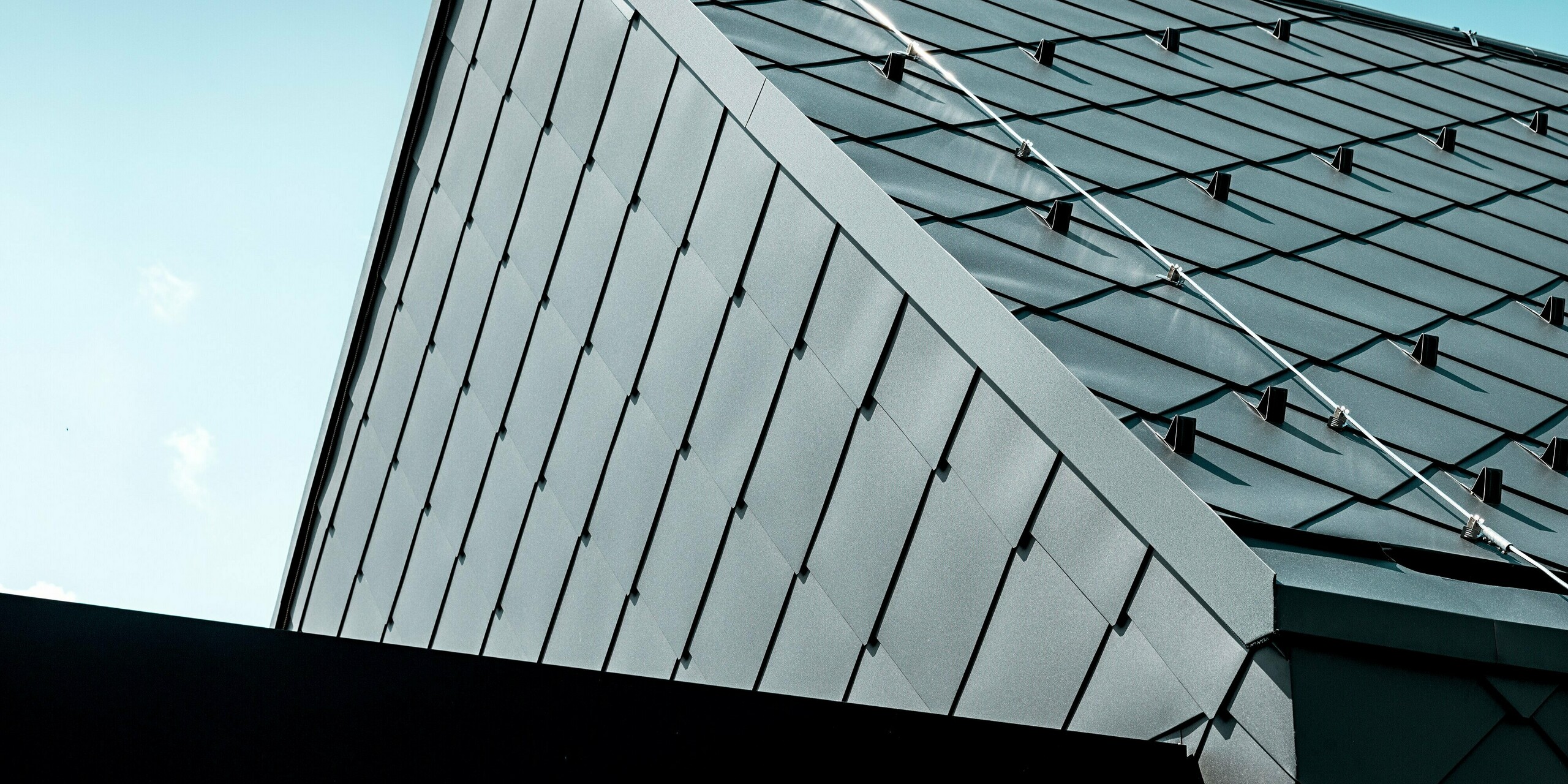 Das Bild zeigt eine markante Detailansicht des Einfamilienhauses in Trnava‑Modranka, dessen Dach vollständig mit PREFA Dachrauten 44 × 44 in P.10 Dunkelgrau gedeckt ist. Die präzise verlegte Rautenstruktur sowie die integrierten Schneestopper betonen die hochwertige Verarbeitung und technische Funktionalität der Aluminiumdeckung. Die matte Oberfläche reflektiert das Licht gleichmäßig und unterstreicht die klare, moderne Form des Giebels. Die langlebigen PREFA Produkte bieten optimalen Schutz, hohe Stabilität und ein zeitlos elegantes Erscheinungsbild.