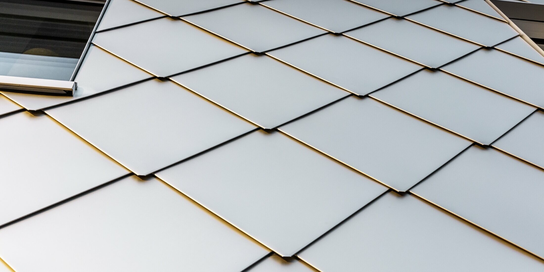 Detailaufnahme der Fassade des Solitaire-Hochhauses in Horw, Luzern, das von der Architektin Tilla Theus entworfen wurde. Die Fassade besteht aus eloxierten PREFA Aluminiumrauten, die die Umgebung reflektieren. Die Rauten aus Blech sind in einem diagonalen Muster angeordnet und verleihen der Oberfläche eine strukturierte, glänzende Optik. Zwei Fenster sind teilweise im Bild sichtbar, was die unregelmäßige Anordnung der Fenster im Gebäude vermuten lässt. 