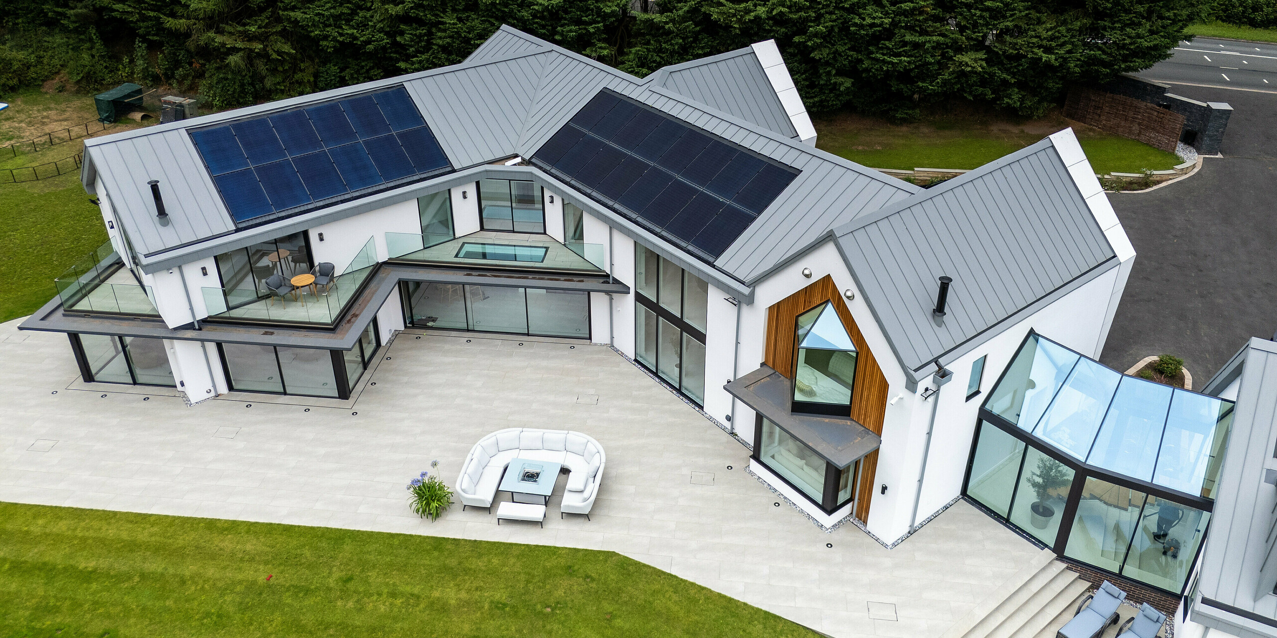 La vue aérienne révèle l'impressionnante villa moderne de Draycott In The Clay, dotée d'une toiture PREFALZ gris quartz. Grâce à sa structure à joints debout épurée et son système photovoltaïque intégré, la toiture allie élégance et haute performance énergétique. La large façade vitrée et le remarquable bardage en bois à l'angle de la maison soulignent la complexité architecturale de la propriété. La vaste terrasse et le jardin paysager créent une harmonie entre le bâtiment et la nature, tandis que la toiture en aluminium PREFALZ garantit une protection et une longévité maximales.