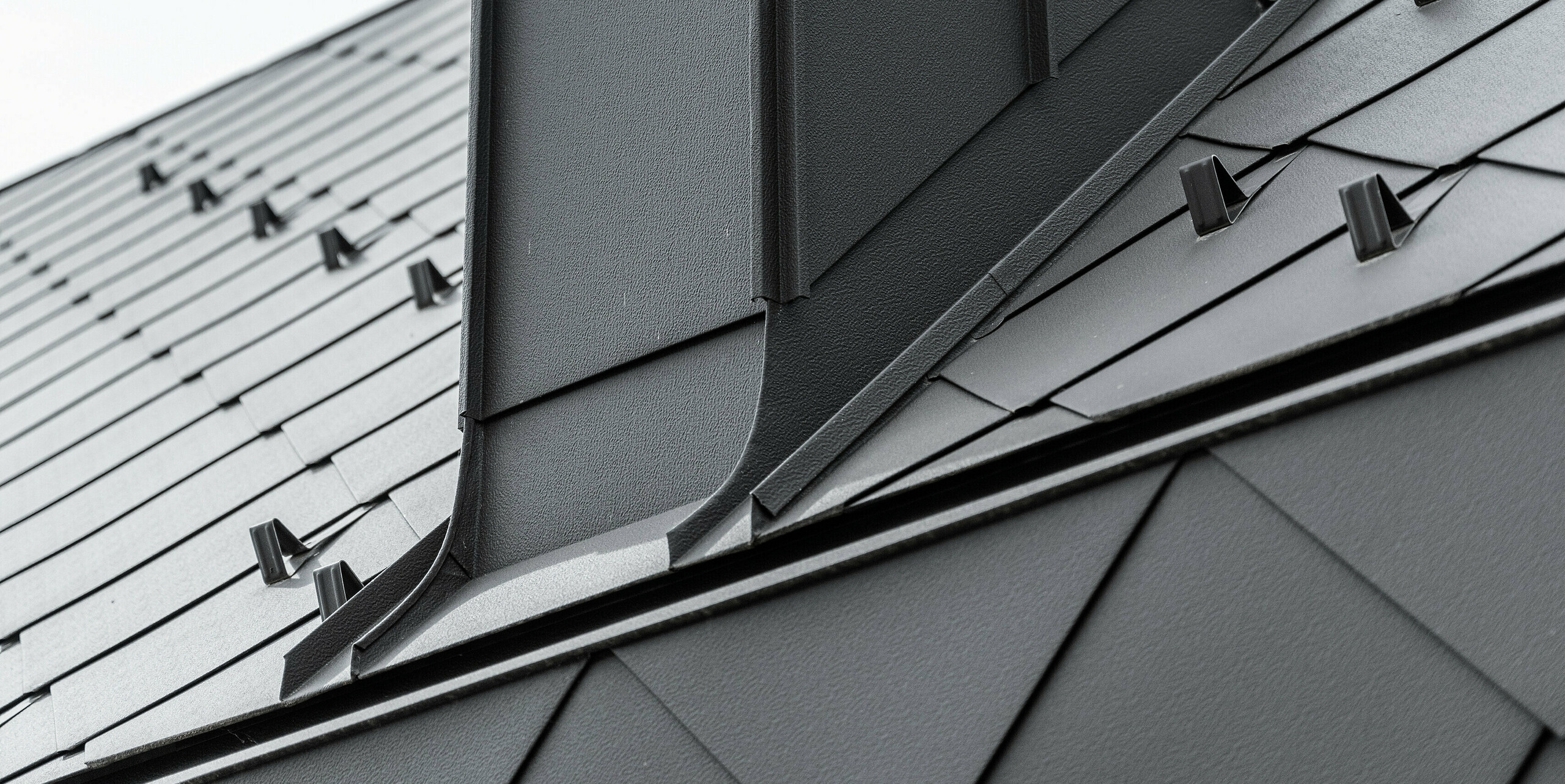Die Traufe eines modernen Reihenhauses in Kaltern, Südtirol, besteht aus geknickten PREFA Dachrauten 44x44. Die Nahaufnahme zeigt die präzise Verarbeitung der robusten und widerstandsfähigen Blechprodukte. Die symmetrische Anordnung der Rauten aus Aluminium und die präzisen Kanten des Schornsteins heben die klaren Linien und die strukturierte Oberfläche hervor, welche die architektonische Eleganz des Gebäudes betont.