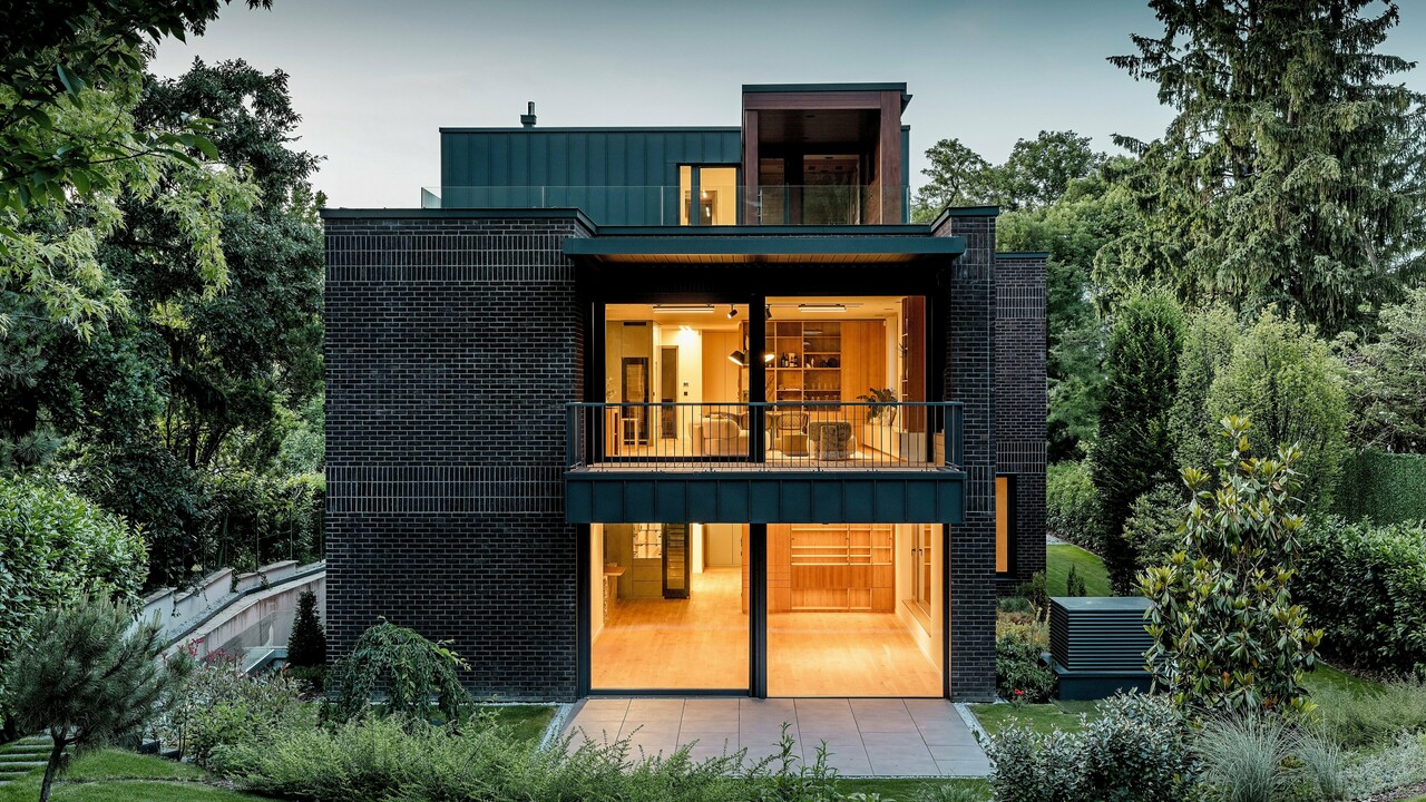 Die Aufnahme zeigt die moderne Rückseite der Budapester Mehrgenerationen‑Villa in der Abenddämmerung. Großzügige Glasfronten öffnen den Blick auf warm erleuchtete Innenräume, während die Kombination aus dunkelroter Backsteinfassade und PREFALZ Aluminium in P.10 Anthrazit klare architektonische Linien betont. Die präzise ausgeführten Winkelstehfalze unterstreichen die hochwertige Verarbeitung und Langlebigkeit der PREFA Aluminiumprodukte. Umgeben von üppigem Grün fügt sich das Gebäude harmonisch in die Gartenlandschaft ein und vermittelt einen eleganten, modernen Charakter.