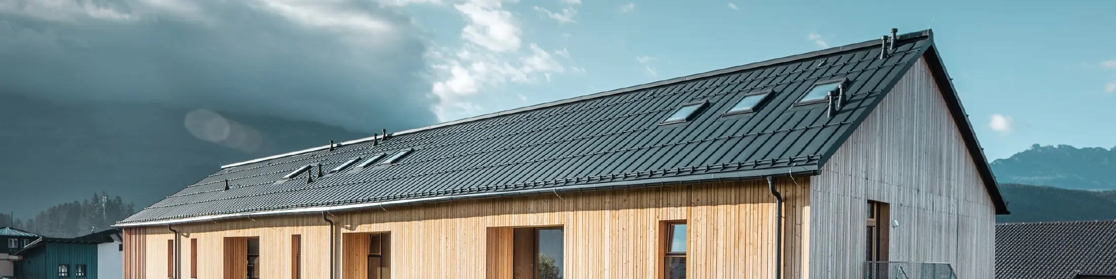 Modernes, langgestrecktes Gebäude mit Satteldach aus dunkelgrauem Aluminium und Fassade aus vertikaler Holzverschalung über weißem Sockel. Große Fensteröffnungen und eine Außentreppe aus Metall betonen die funktionale Architektur. Das Gebäude steht auf einer grünen Wiese mit umliegenden Häusern und Bergen im Hintergrund unter teils wolkigem Himmel.