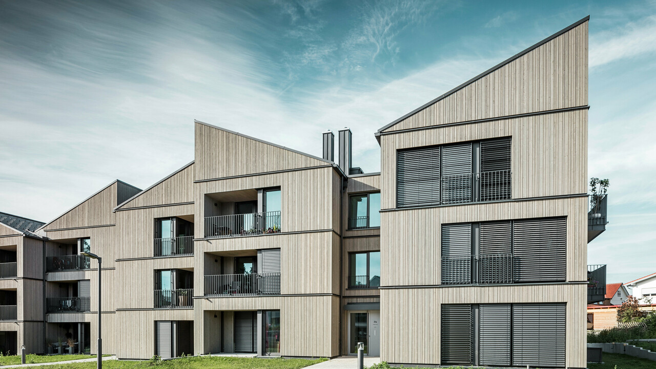 Zone d'entrée d'un immeuble du quartier Schmucker à Utting am Ammersee. L'architecture impressionne par ses lignes claires et ses toits en appentis frappants, recouverts de R.16 PREFA durables en gris sombre P.10. L'élégante façade en bois s'harmonise parfaitement avec les grandes fenêtres et les balcons qui apportent beaucoup de lumière et une atmosphère conviviale. La conception durable des produits en aluminium PREFA souligne la construction hybride innovante de ce projet résidentiel à l'architecture sophistiquée.