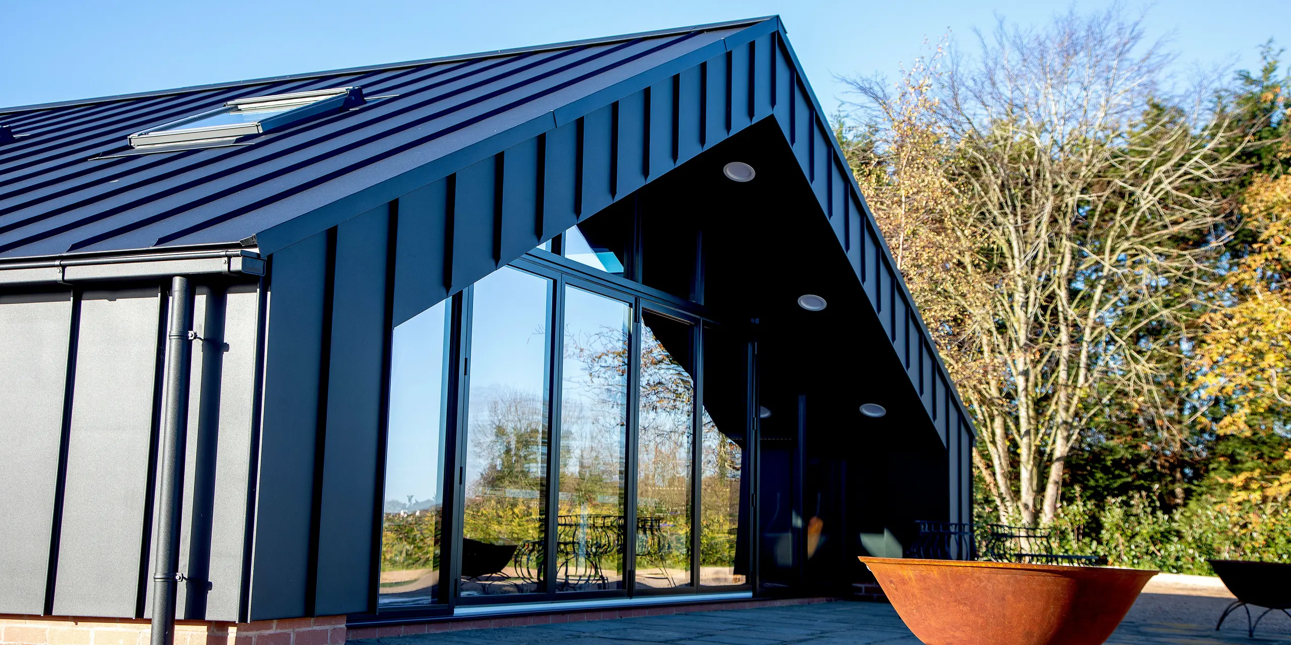 Vue plongeante sur un bâtiment de loisirs moderne, revêtu d'une façade en aluminium PREFA et d'une toiture PREFALZ P.10 Noir. Les lignes de joints debout saisissantes, la géométrie épurée de la toiture et la généreuse façade vitrée confèrent au bâtiment une présence architecturale affirmée. Sa structure en aluminium résistant aux intempéries garantit une stabilité maximale et une protection durable contre les agressions extérieures.