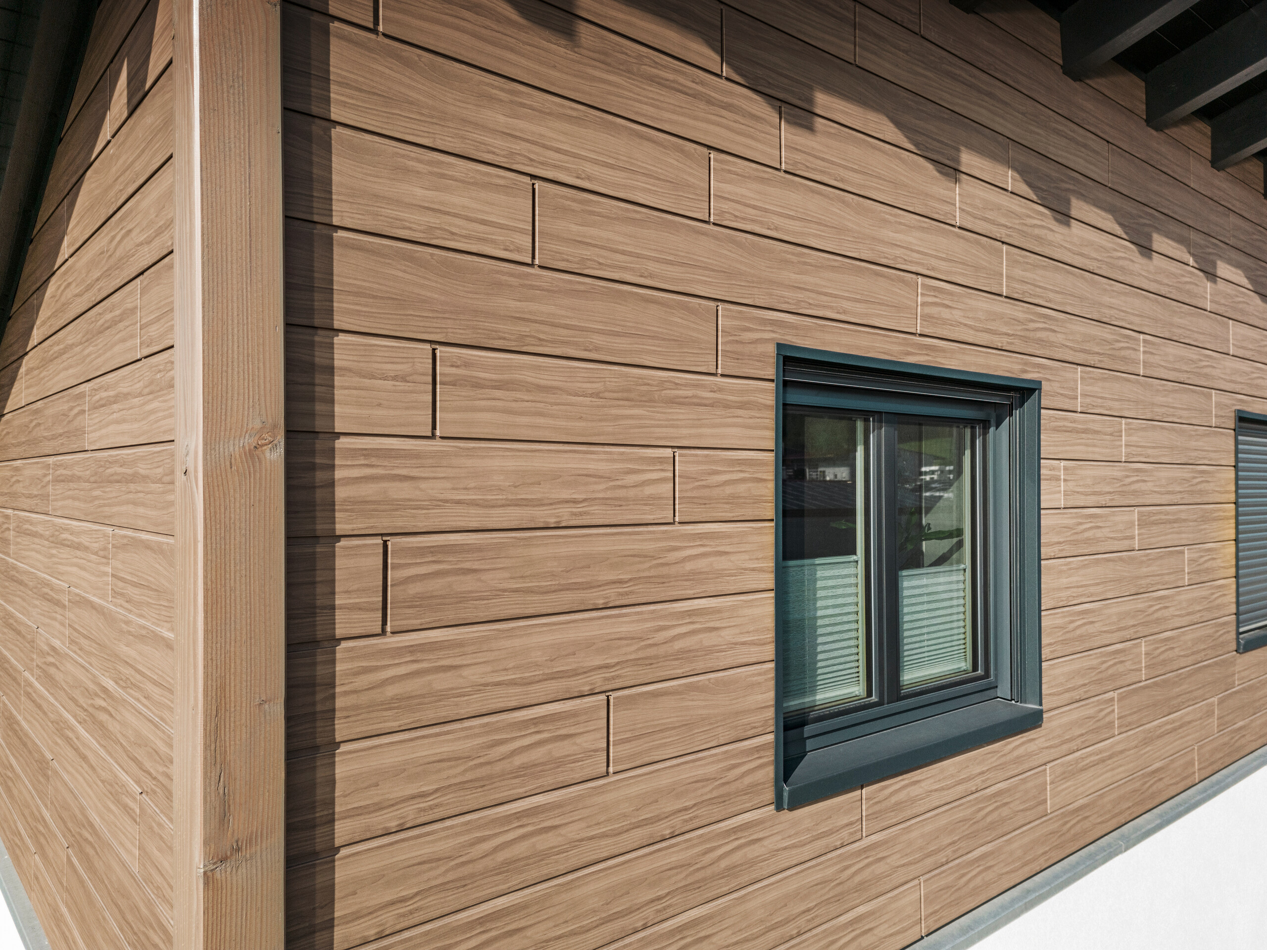 Detailaufnahme der Gebäudekante am Obergeschoss eines Einfamilienhauses in Bruck. Das obere Stockwerk ist mit einer Fassade aus Aluminium in einer Holzoptik verkleidet. Die PREFA Sidings in Walnuss Braun bilden einen harmonischen Übergang zum weißen Gebäudesockel.