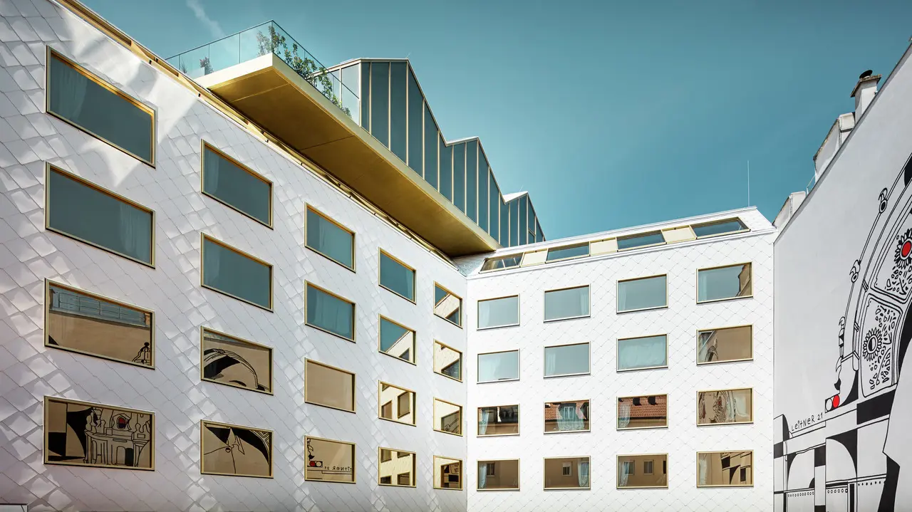 Vista prospettica dell'hotel "THE ROCK Radisson RED Vienna" di Vienna, caratterizzato dall'innovativa facciata in alluminio PREFA con tetto e pareti a rombi 44 × 44 in P.10 bianco puro. Le finestre disposte in modo irregolare con vetri a specchio riflettono dinamicamente l'ambiente circostante.