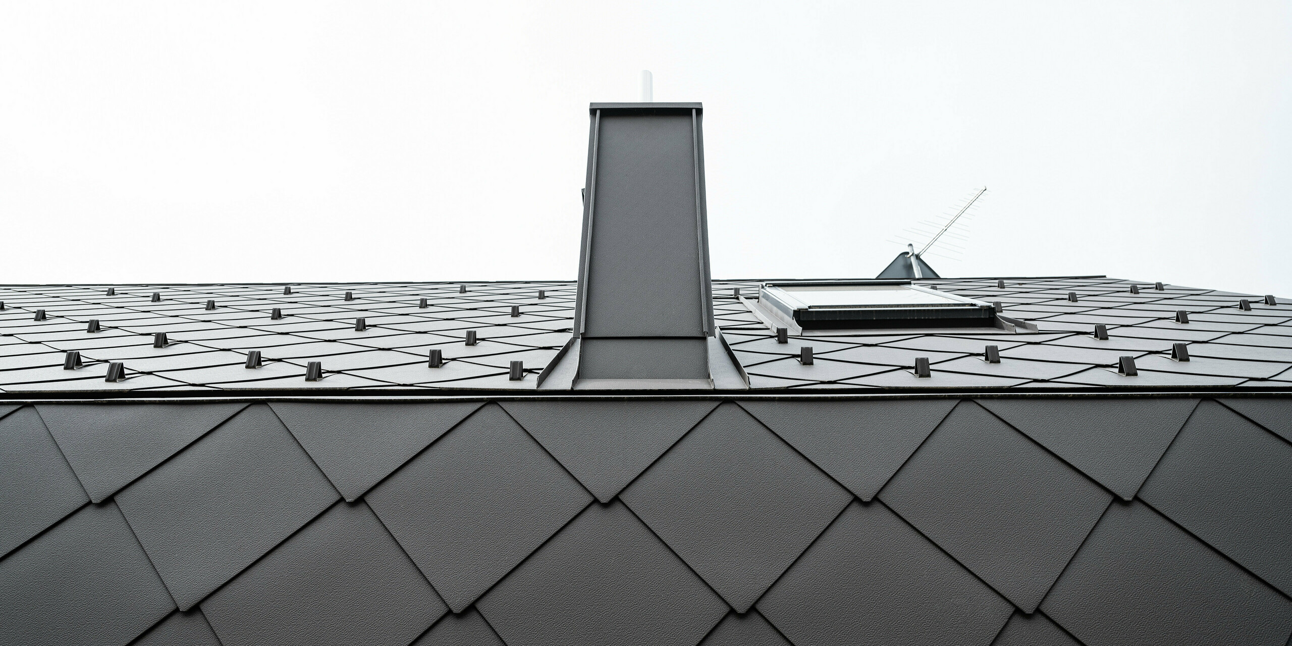 Detailansicht eines modernen Daches aus Blech. Das Gebäude ist ausgestattet mit PREFA Dach- und Wandrauten 44x44 in der Farbe P.10 Braun. in Anthrazit. Die Aufnahme zeigt die präzise Verlegung der Aluminiumrauten, die durch ihre einzigartige Form charakterisiert sind. Zentral im Bild ist ein schmaler Schornstein aus Aluminium zu sehen. An der Traufe wurden die Rauten geknickt, was für eine homogene Gebäudehülle sorgt.