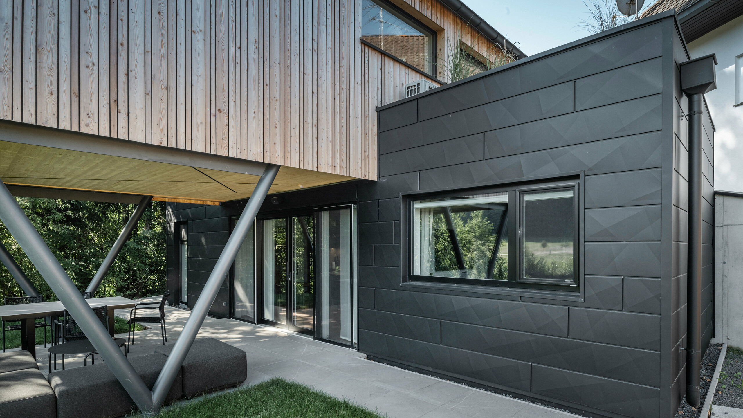Punto di forza architettonico: “Woodstock Chalet 5” a St. Martin am Innkreis, Alta Austria. La moderna struttura ricettiva per matrimoni è valorizzata dall'uso di pannelli per facciate PREFA Siding.X color antracite. Gli elementi in alluminio creano un forte contrasto con i pannelli in legno chiaro e le strutture di sostegno metalliche. I telai delle finestre scuri si integrano armoniosamente nel design della facciata, conferendo all'edificio un aspetto elegante. Questa immagine mostra l'equilibrio estetico tra la texture naturale del legno e il fascino industriale della facciata in alluminio, che rappresenta il design degli edifici contemporanei.