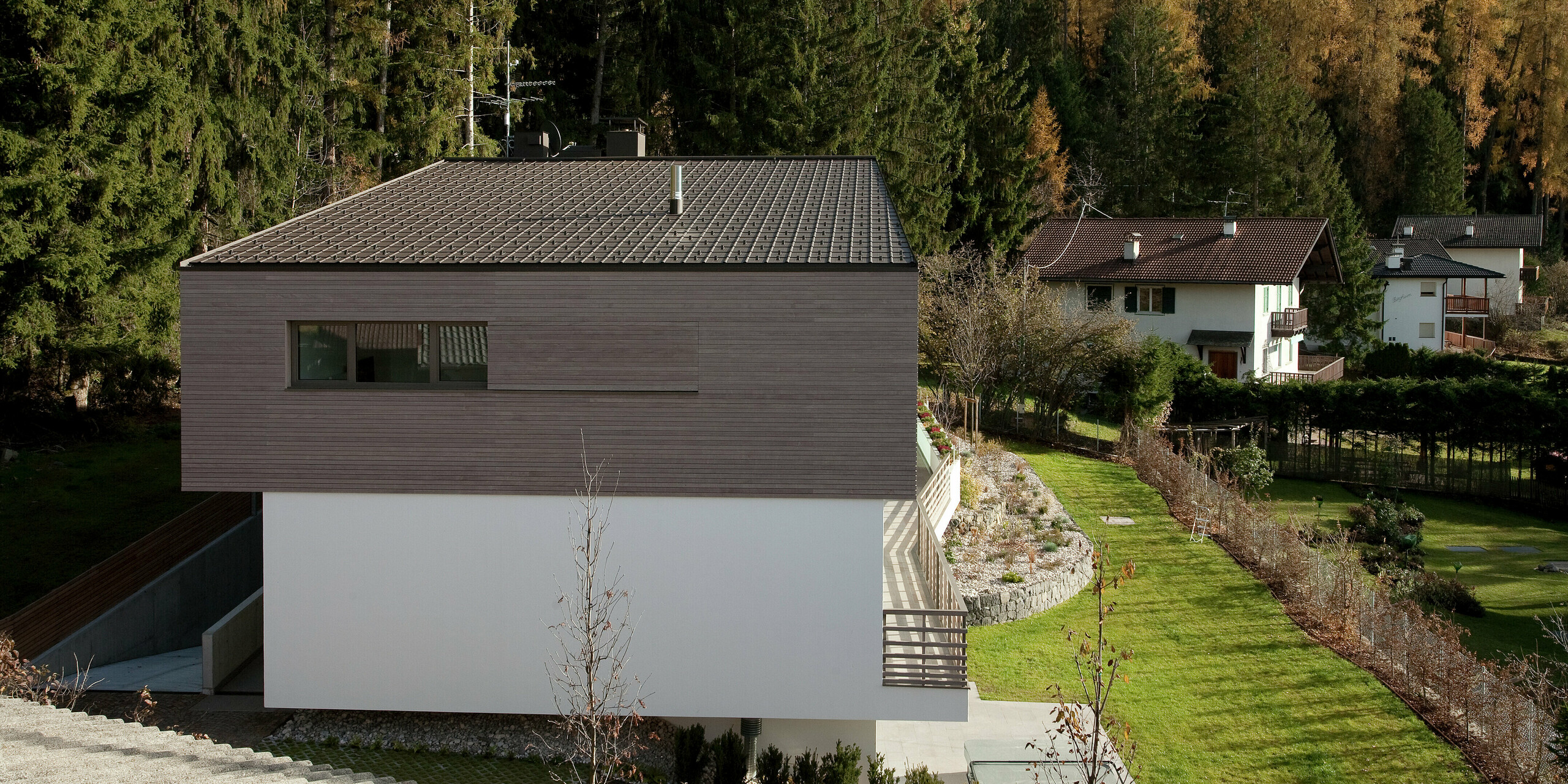 Seitliche Ansicht des Hauses mit PREFA Dachplatten in P.10 Braun und einer großzügigen Terrasse, die den Blick auf die umgebende Natur freigibt. Die Kombination aus Holzfassade und modernen Materialien schafft eine edle Optik.